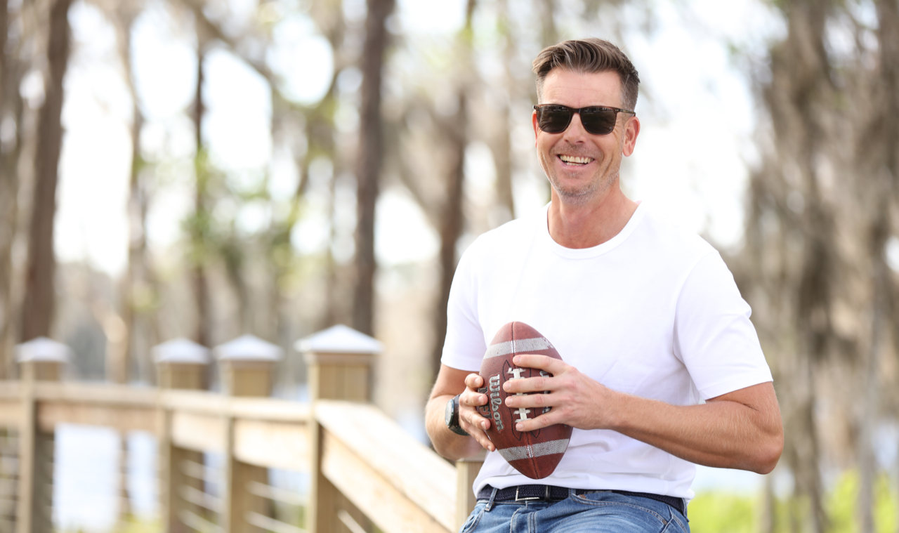 Man with a white t-shirt and sun glasses is sitting on a fence, holding an American football in his hands.