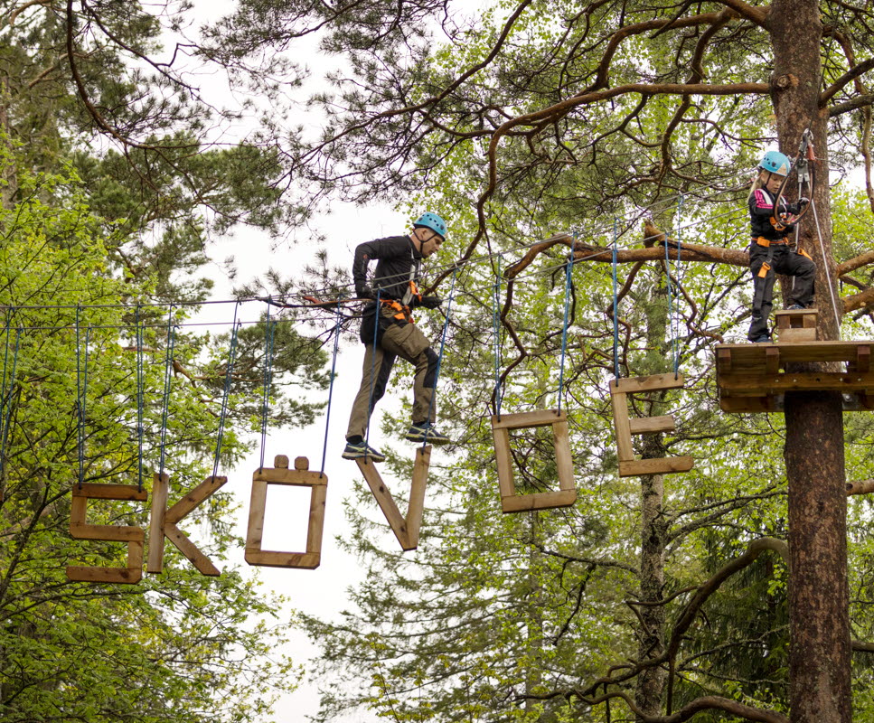 En man klättrar i höghöjdsbana i skog på bokstäver som bildar ordet Skövde. 