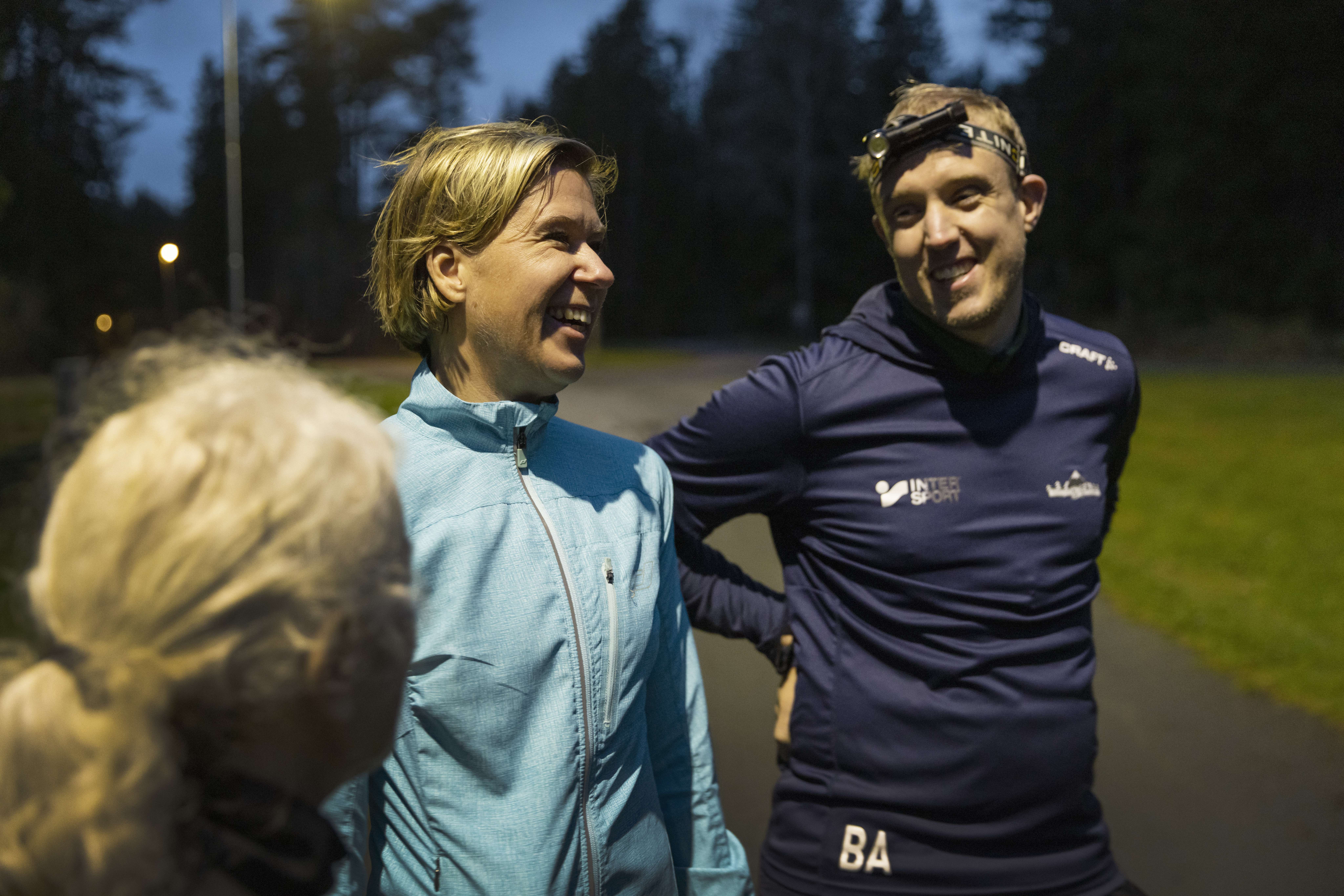 Tre löparklädda personer ler glatt mot varandra mitt i ett samtal. En har en pannlampa på sig. 