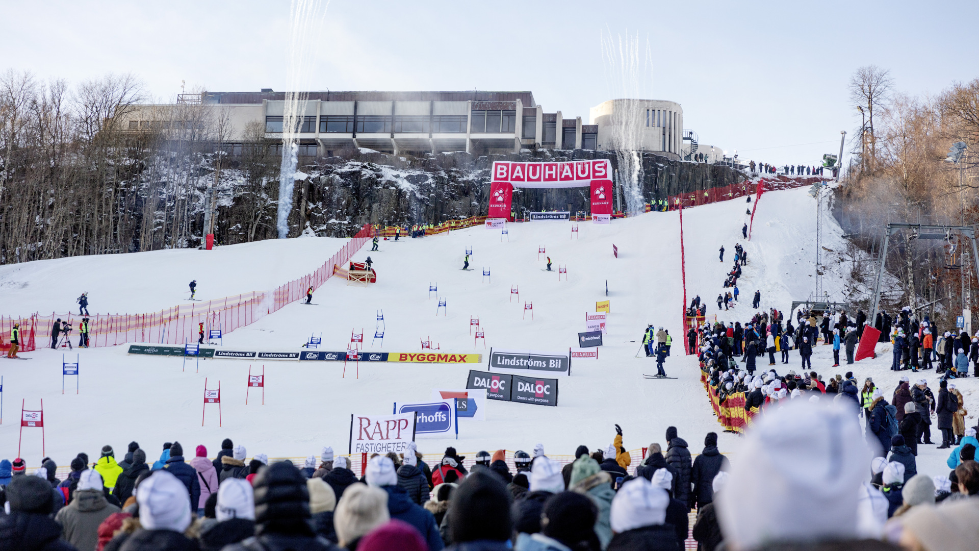 Människor som står vid målgången i en slalomtävling. I bakgrunden syns startbågen och alla flaggor längs backen.