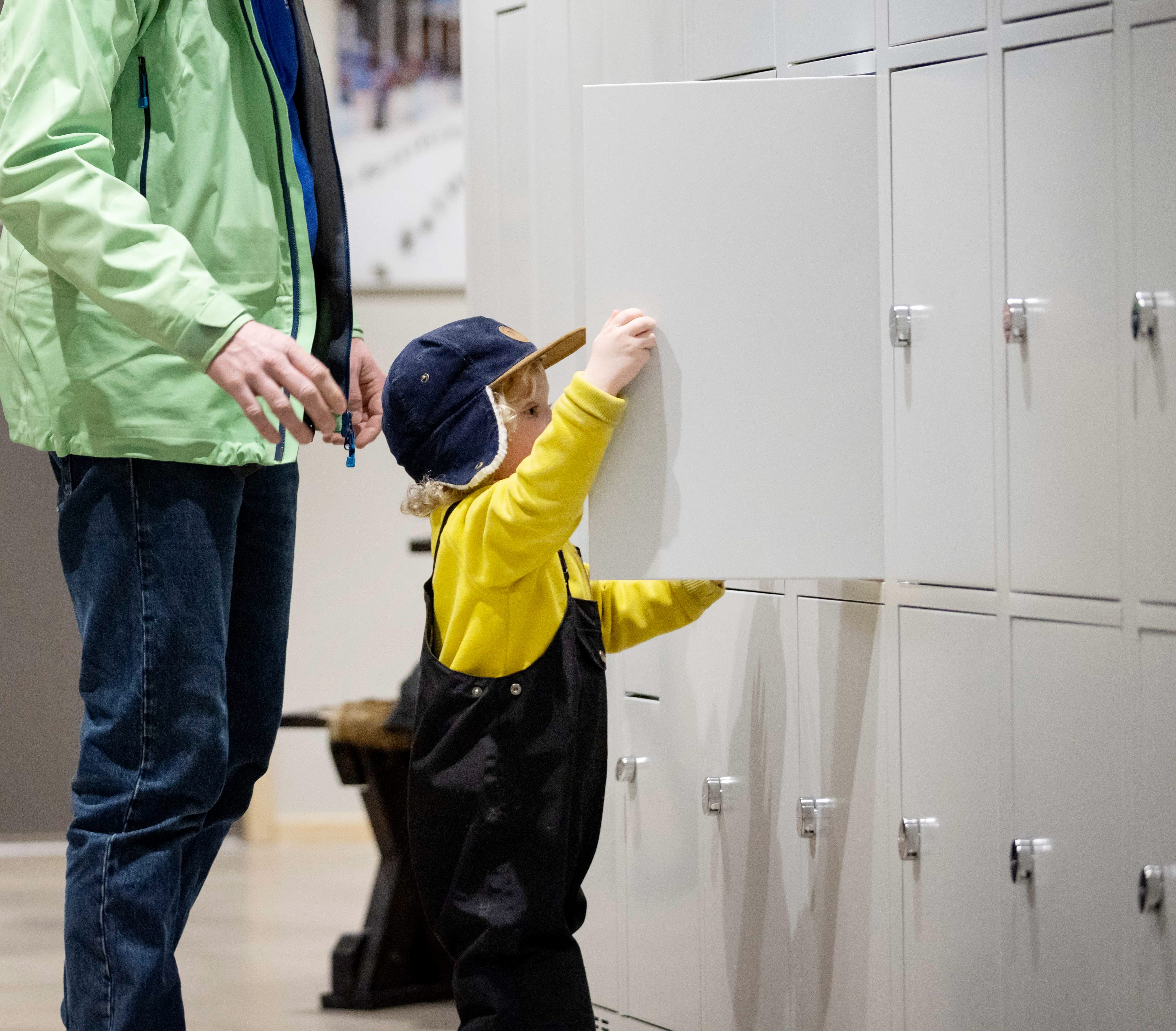 En vägg är täckt av förvaringsskåp. En liten kille står och stänger ett av skåpen. Bakom honom står hans pappa. 