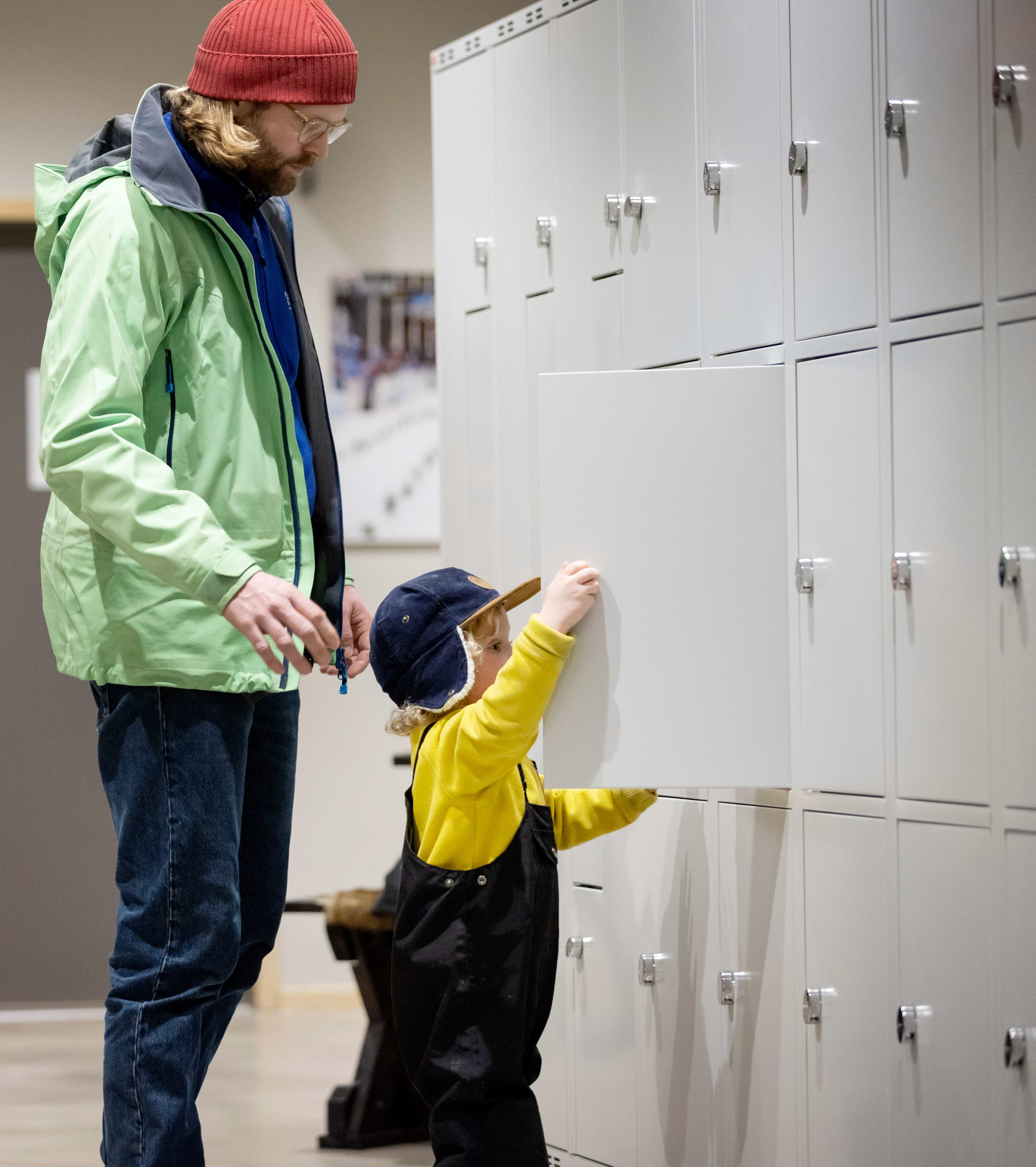 En vägg är täckt av förvaringsskåp. En liten kille står och stänger ett av skåpen. Bakom honom står hans pappa. 