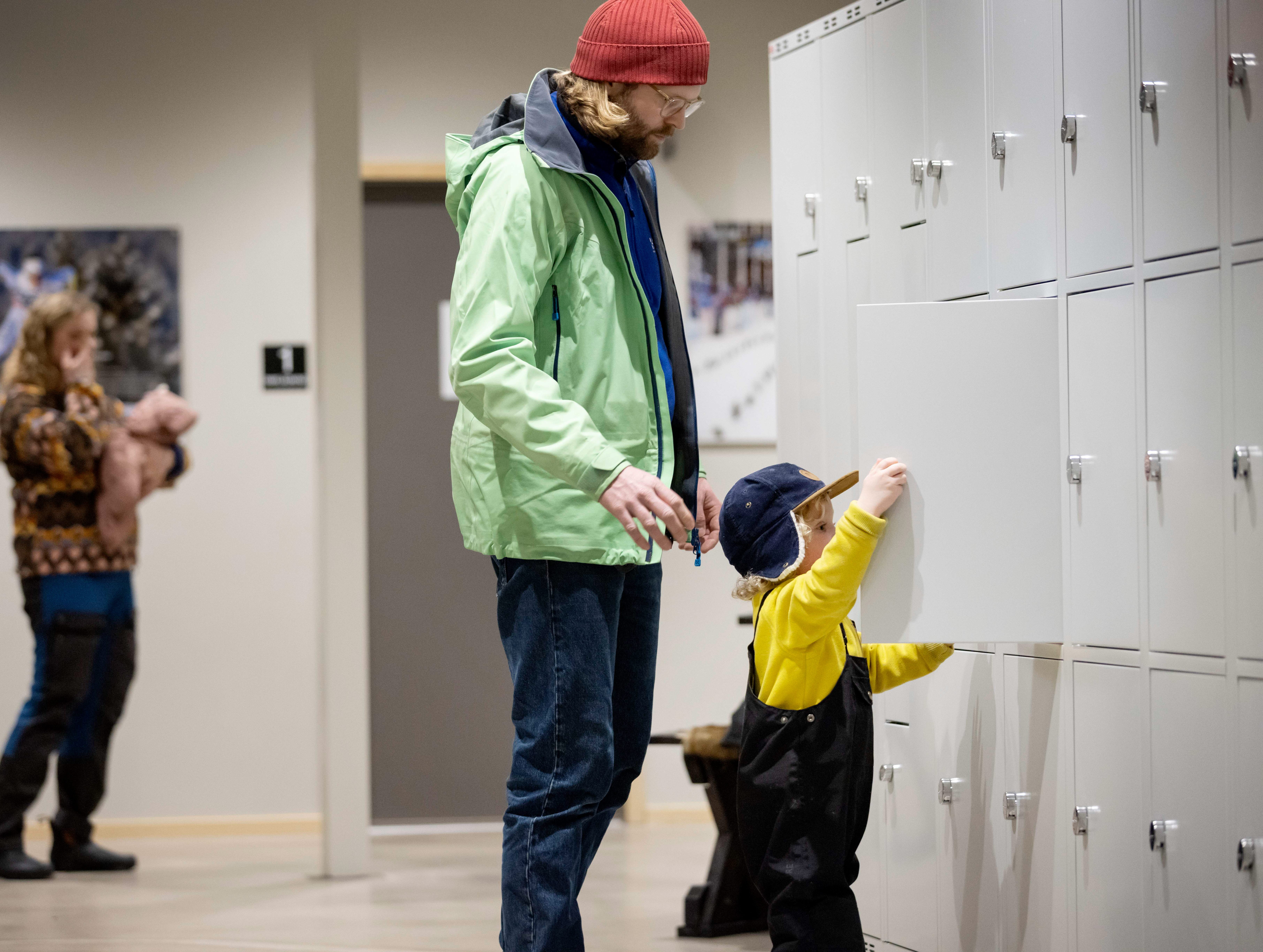En vägg är täckt av förvaringsskåp. En liten kille står och stänger ett av skåpen. Bakom honom står hans pappa. 