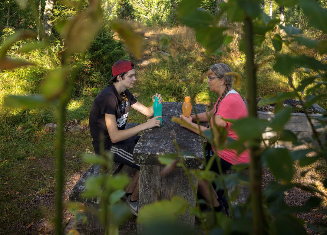 Mor och son sitter vid picknickbord vid Ymsingsborg på Billingen Skövde, grenar skymmer dem något i förgrunden. 