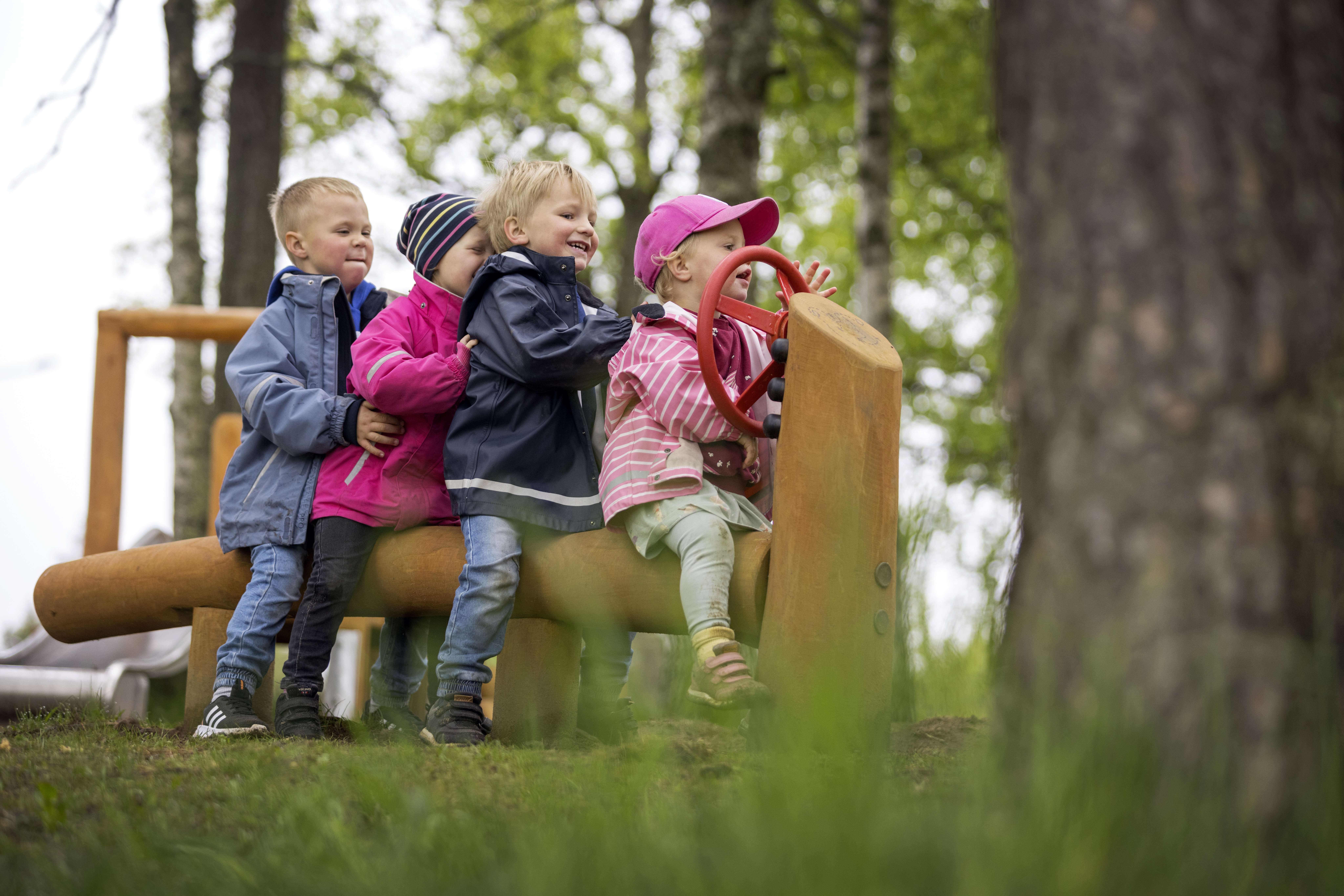 Four small children sit behind each other on a long wooden log. The child at the front has a steering wheel on a wooden post in front of him.
