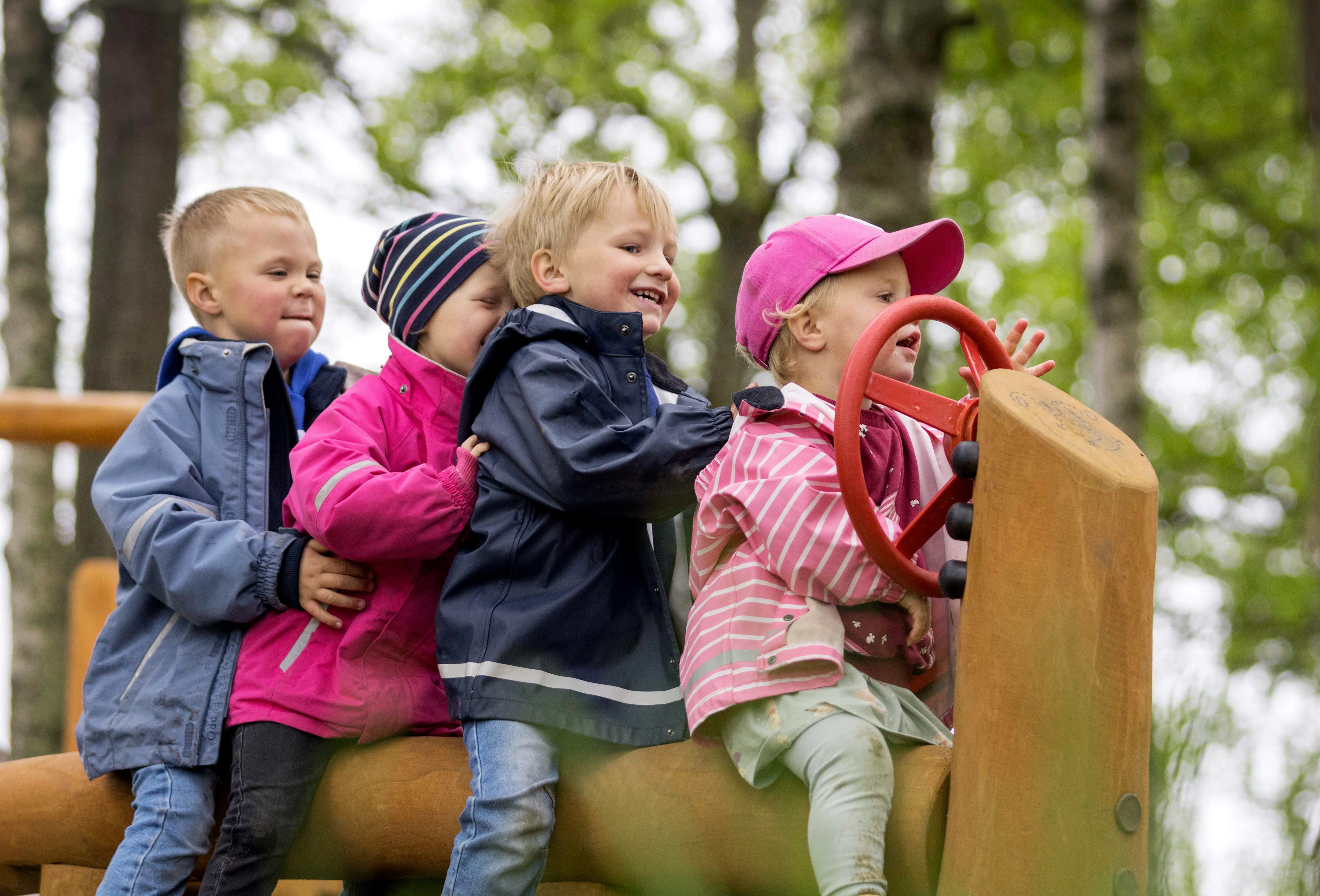 Fyra små barn sitter bakom varandra på en lång trästock. Barnet längst fram har en ratt på en trästolpe framför sig. 