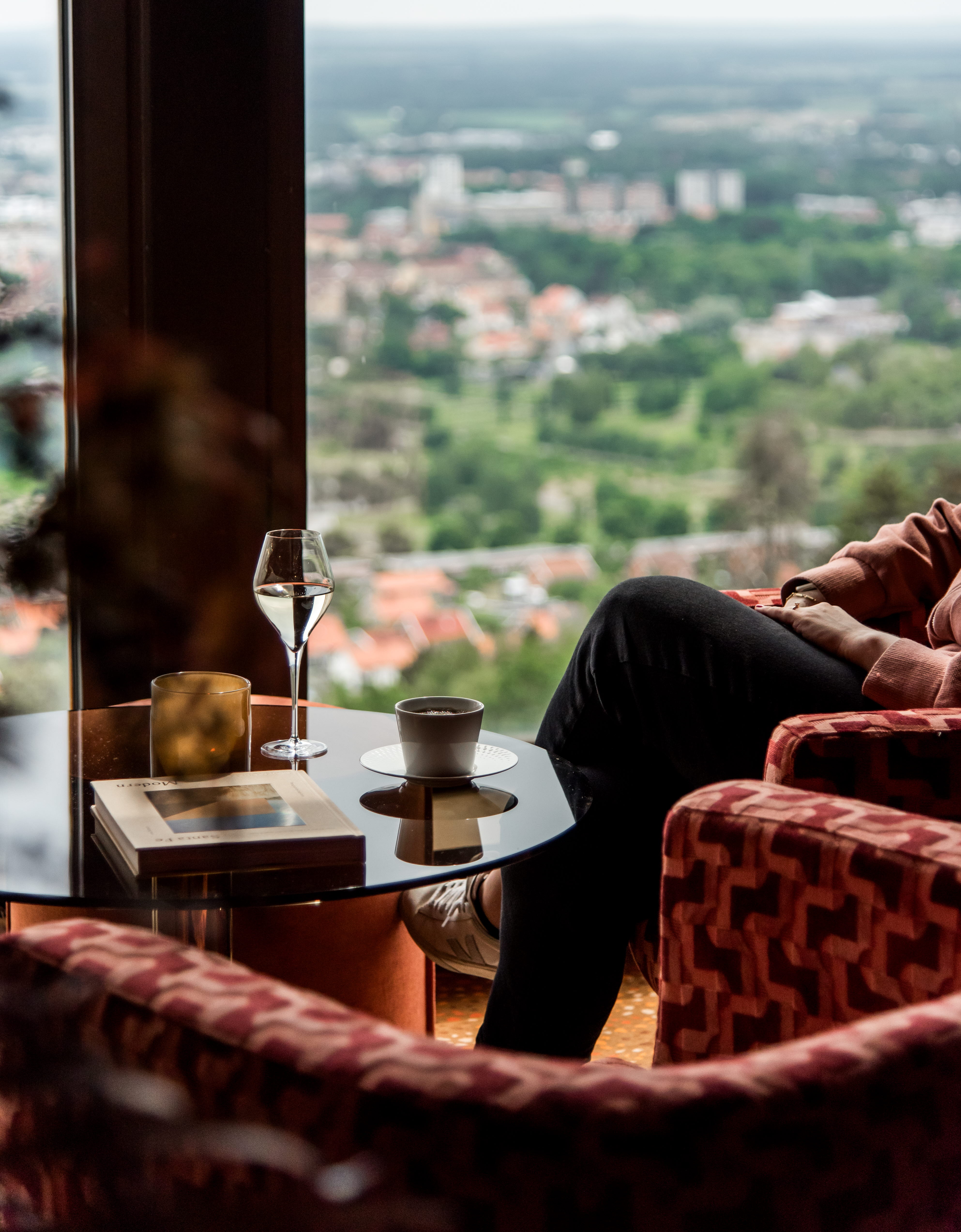 En person sitter i en röd fåtölj med ett glas champagne på ett glasbord framför sig. I bakgrunden ser man en utsikt över Skövde.