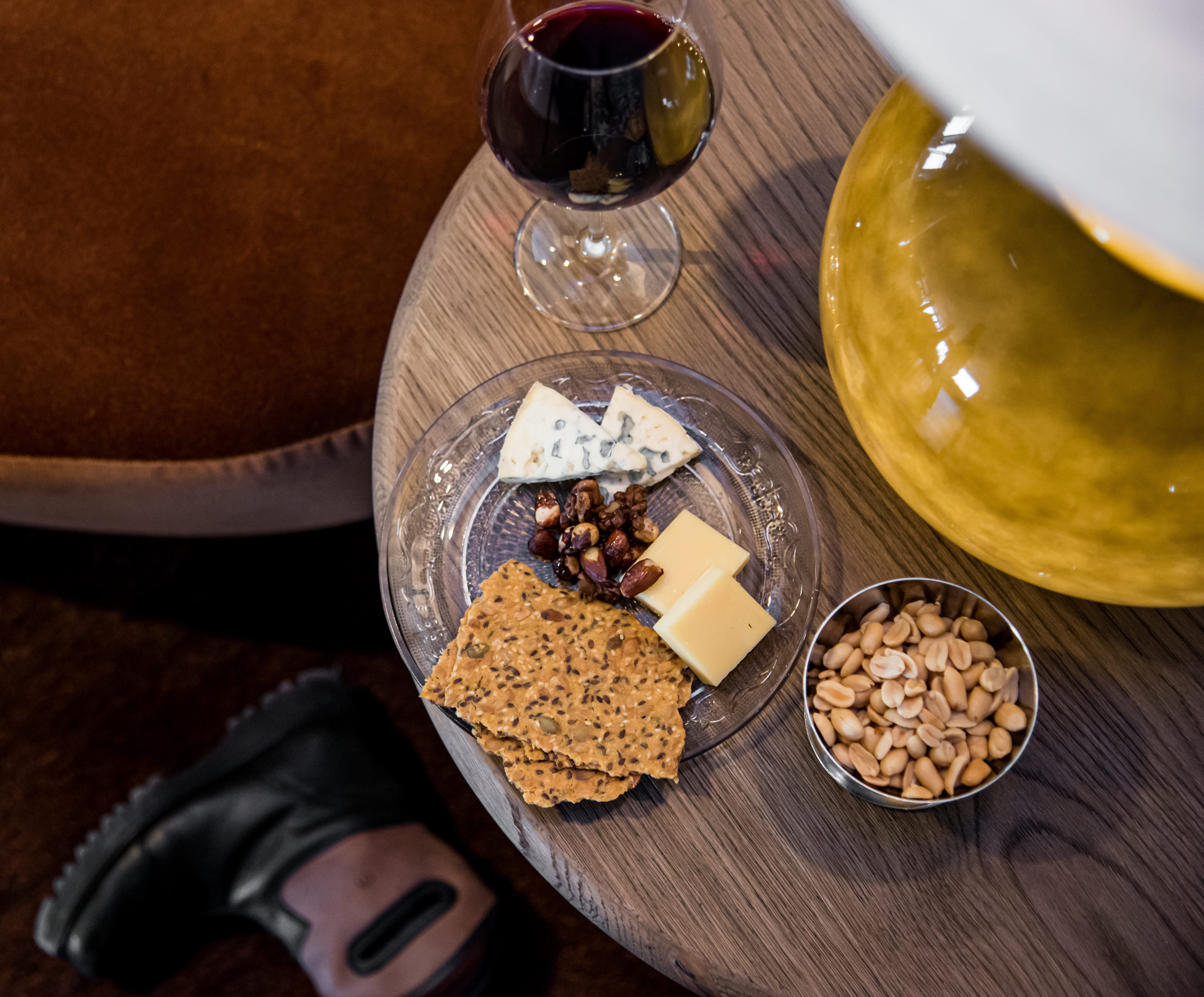 A picture of a wooden table with a glas of red wine, cheese and some peanuts. 