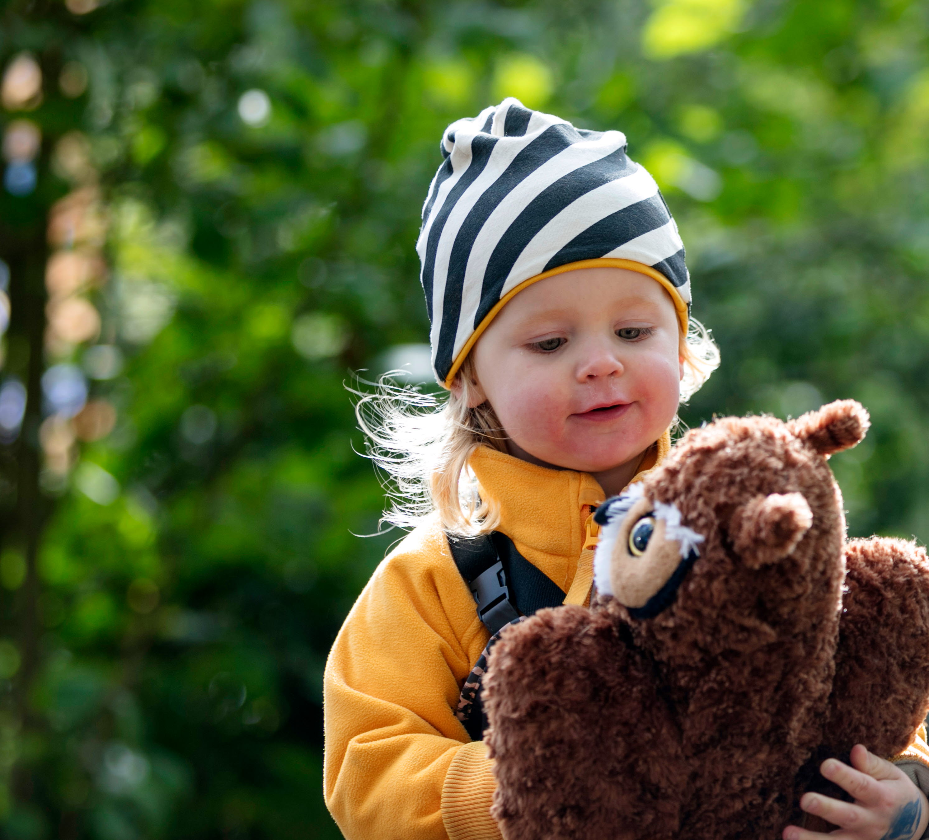 Flicka i gul tröja och svart/vit randig mössa leker med en nalle tillsammans med Skogsmulle.
