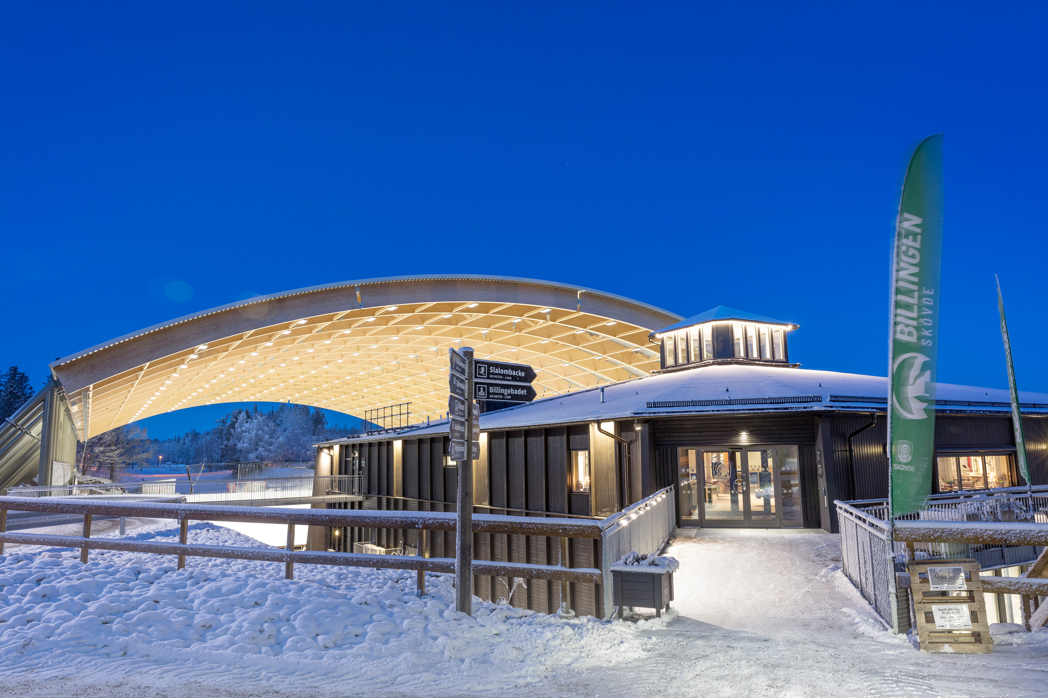 Upplyst svart byggnad(Billingecenter) i skymningen. Det liger snö på backen och i bakgrunden ser man det välvda taket över isbanan. 