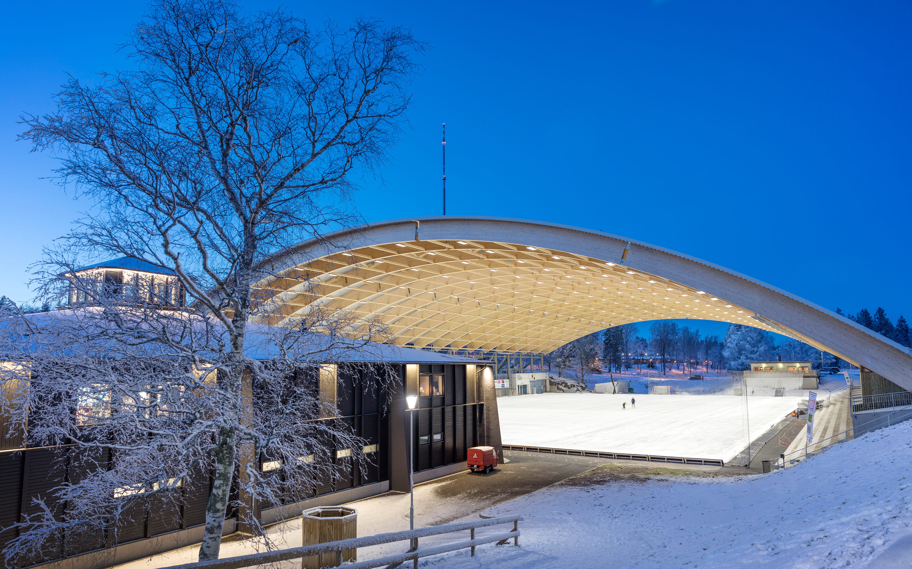 Upplyst svart byggnad(Billingecenter) i skymningen. Det liger snö på backen och i bakgrunden ser man det välvda taket över isbanan. 