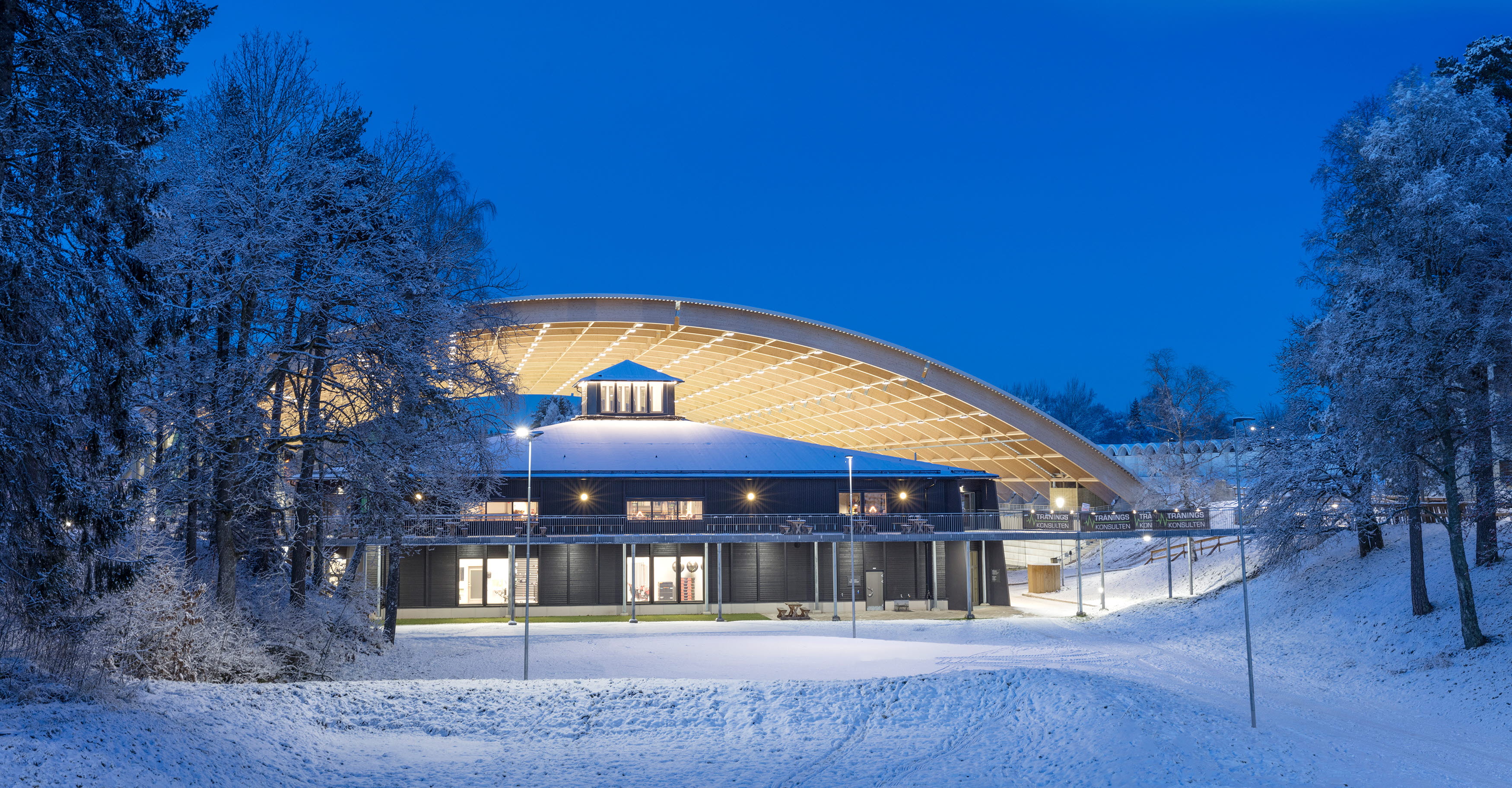 Upplyst svart byggnad(Billingecenter) i skymningen. Det liger snö på backen och i bakgrunden ser man det välvda taket över isbanan. 