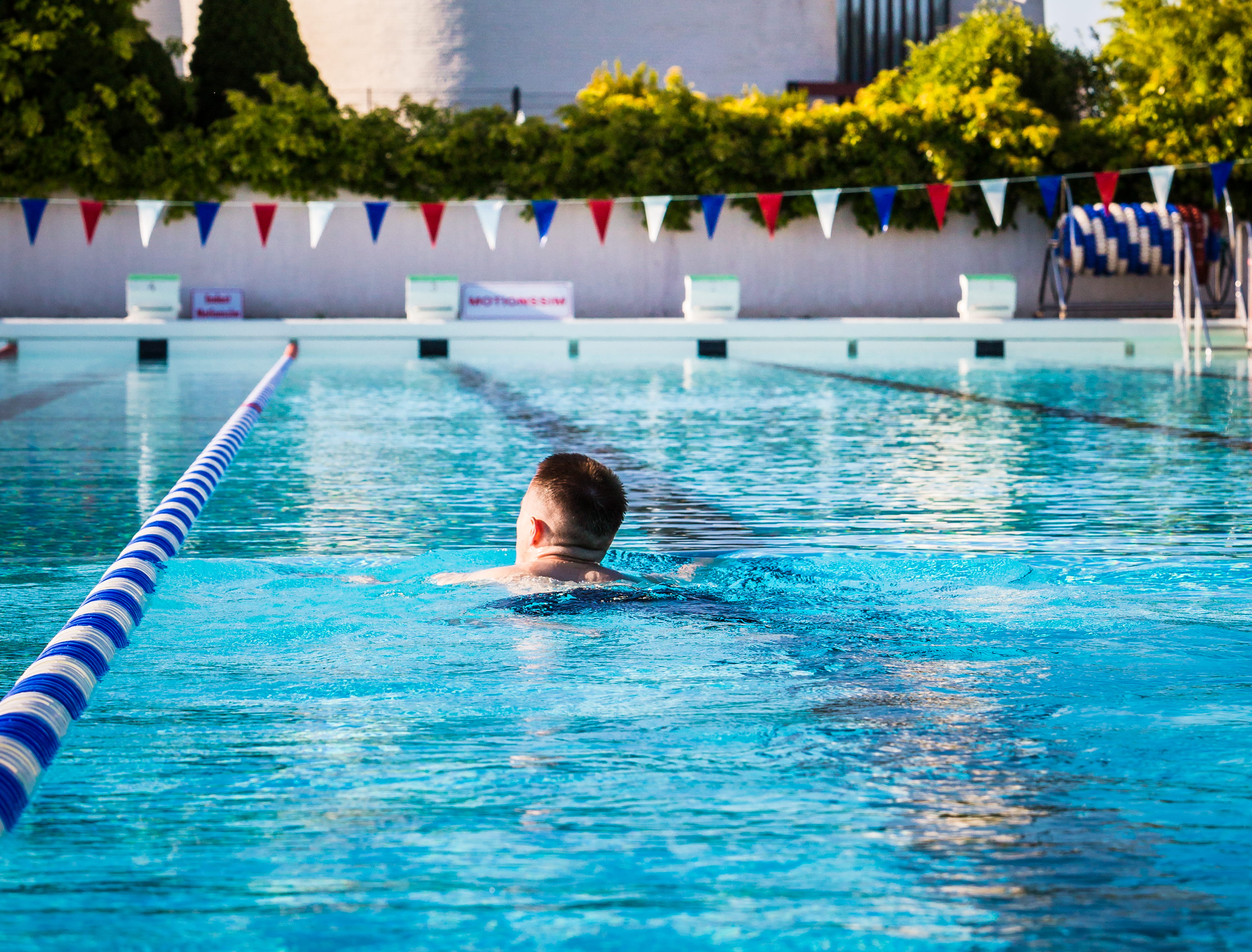 En tidig sommarmorgon vid Billingebadet. Över bassängens turkosa vatten hänger flaggspel. I bassängen simmar en motionssimmare. I bakgrunden syns de vita, runda väggarna från Sankt Lukas kyrka. 