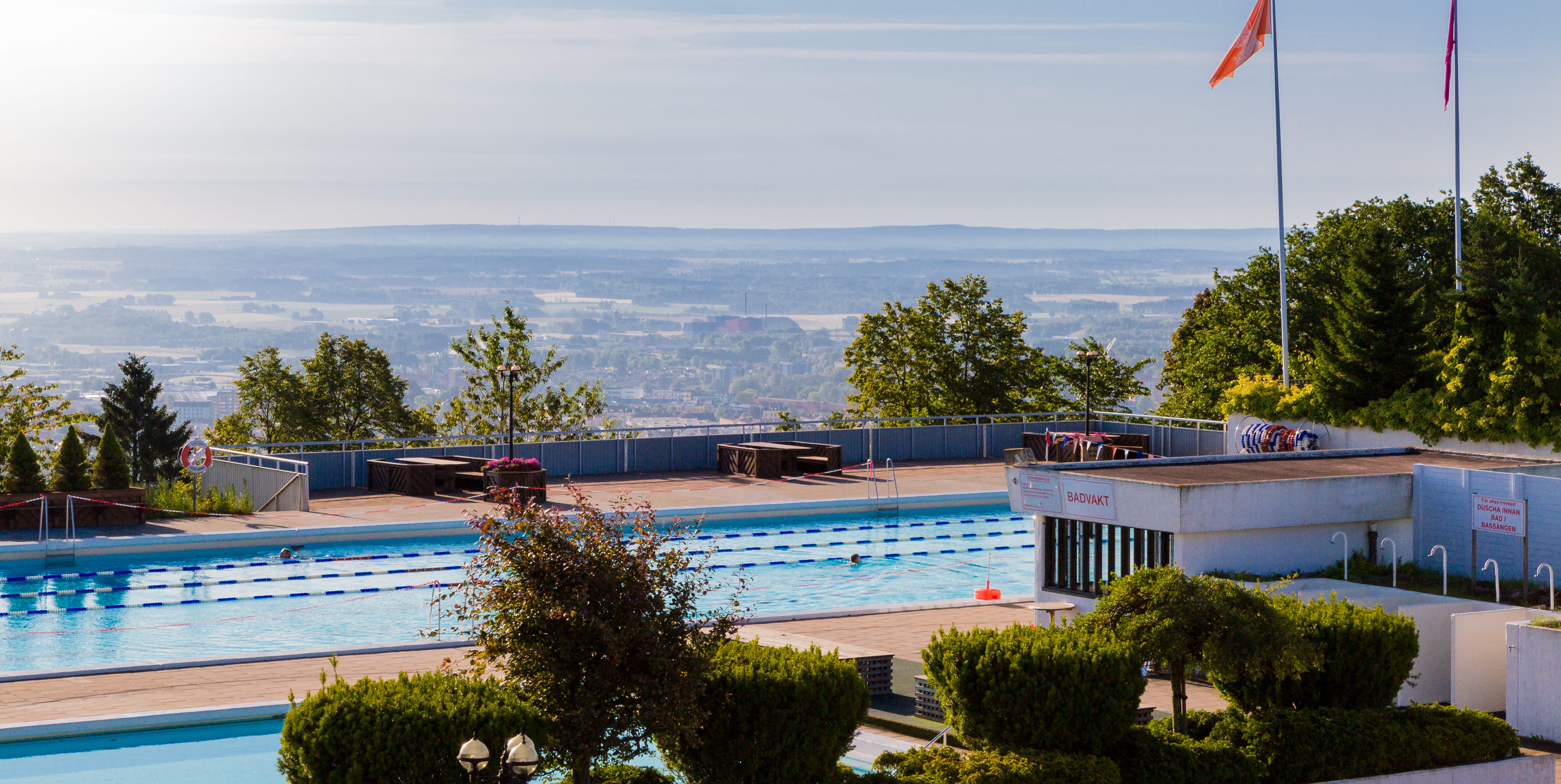 En tidig sommarmorgon vid Billingebadet. Strålande solsken på klarblå himmel, med bara små duntussar till moln som moln. Bassängerna ligger vid kanten av berget Billingen, nedanför ser man staden Skövde.