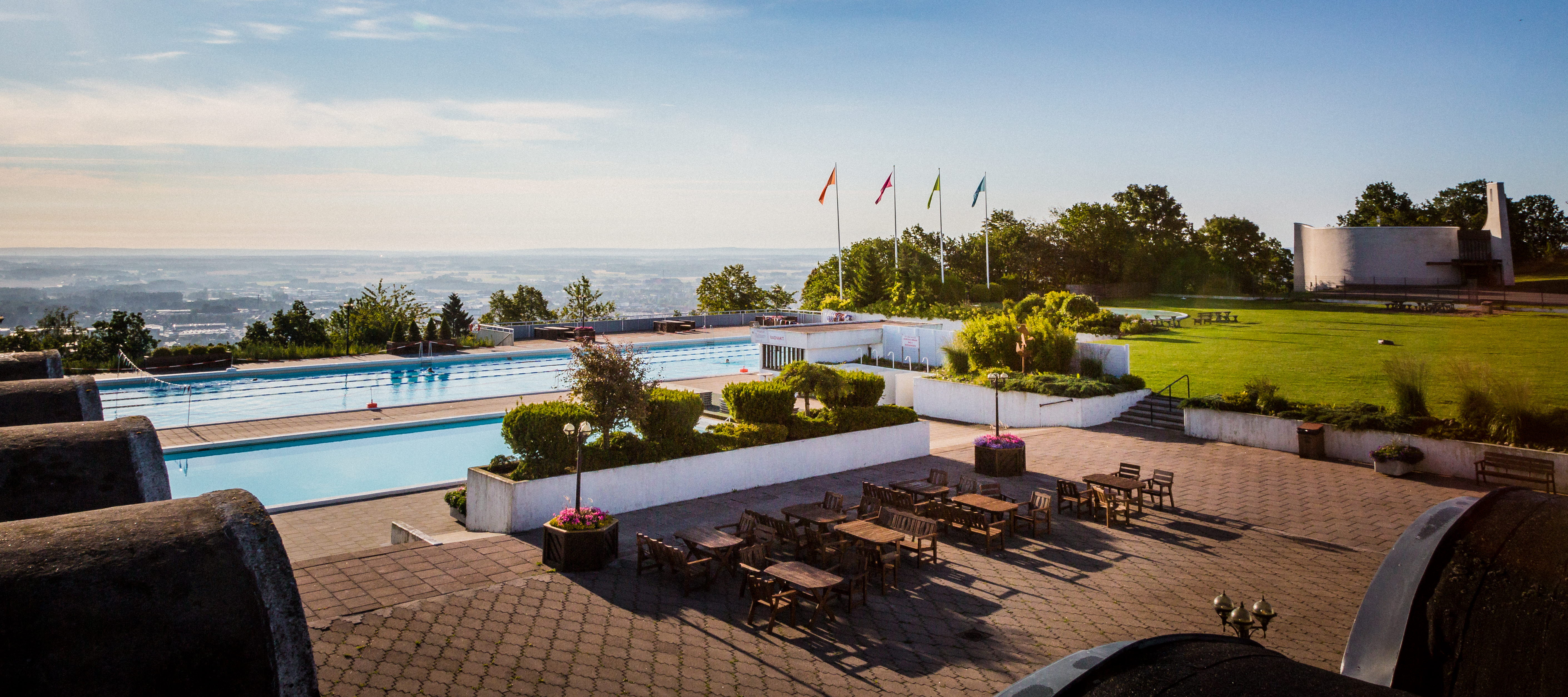 En tidig sommarmorgon vid Billingebadet. Strålande solsken på klarblå himmel, med bara små duntussar till moln som moln. Bassängerna ligger vid kanten av berget Billingen, nedanför ser man staden Skövde.