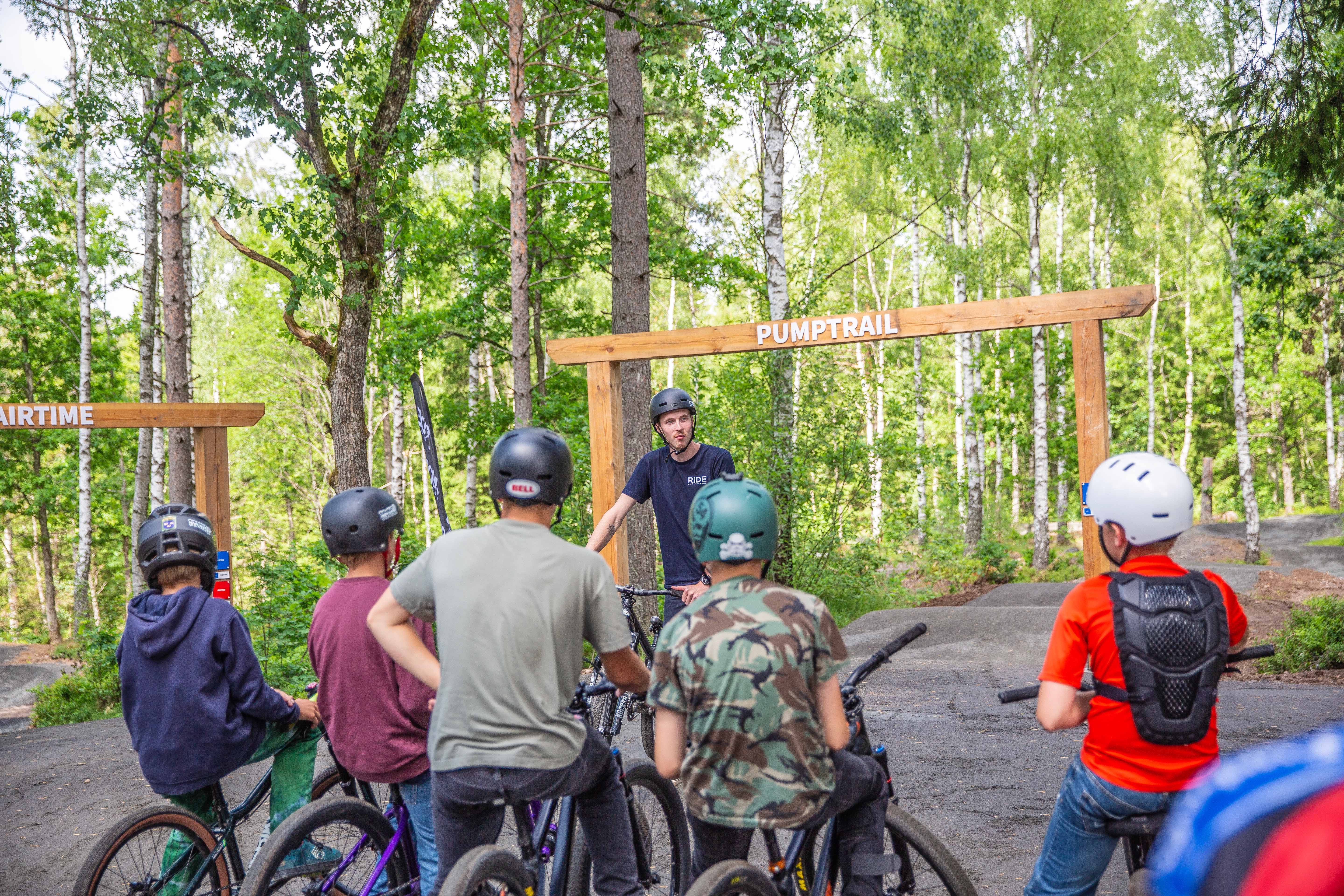 A person in a dark t-shirt and black helmet  talking in front of five children who stand ready with their bikes in front of a mtb arena.