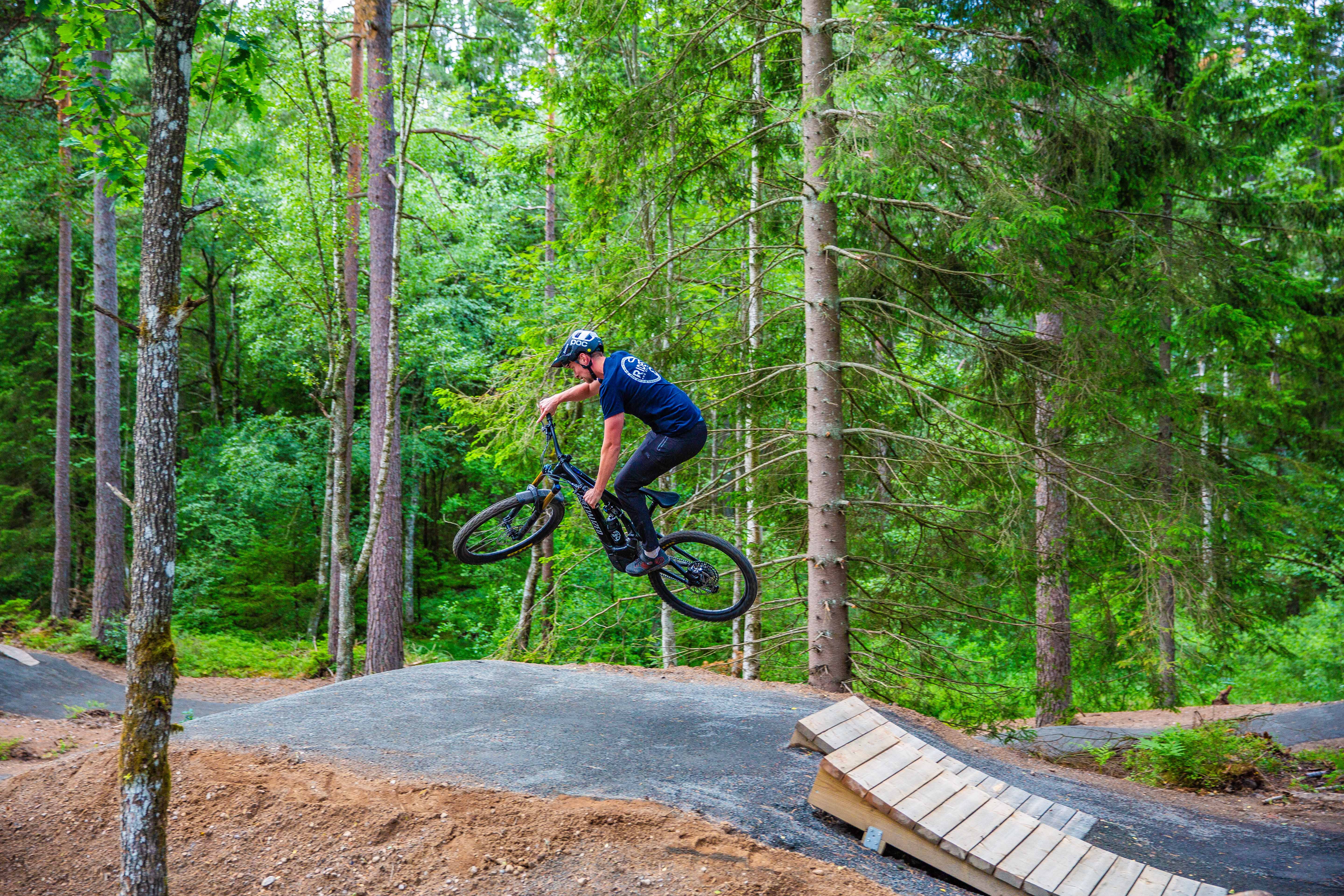 A person in dark clothes and a helmet jumps into the air from a wooden ramp with a mtb bike in the forest.