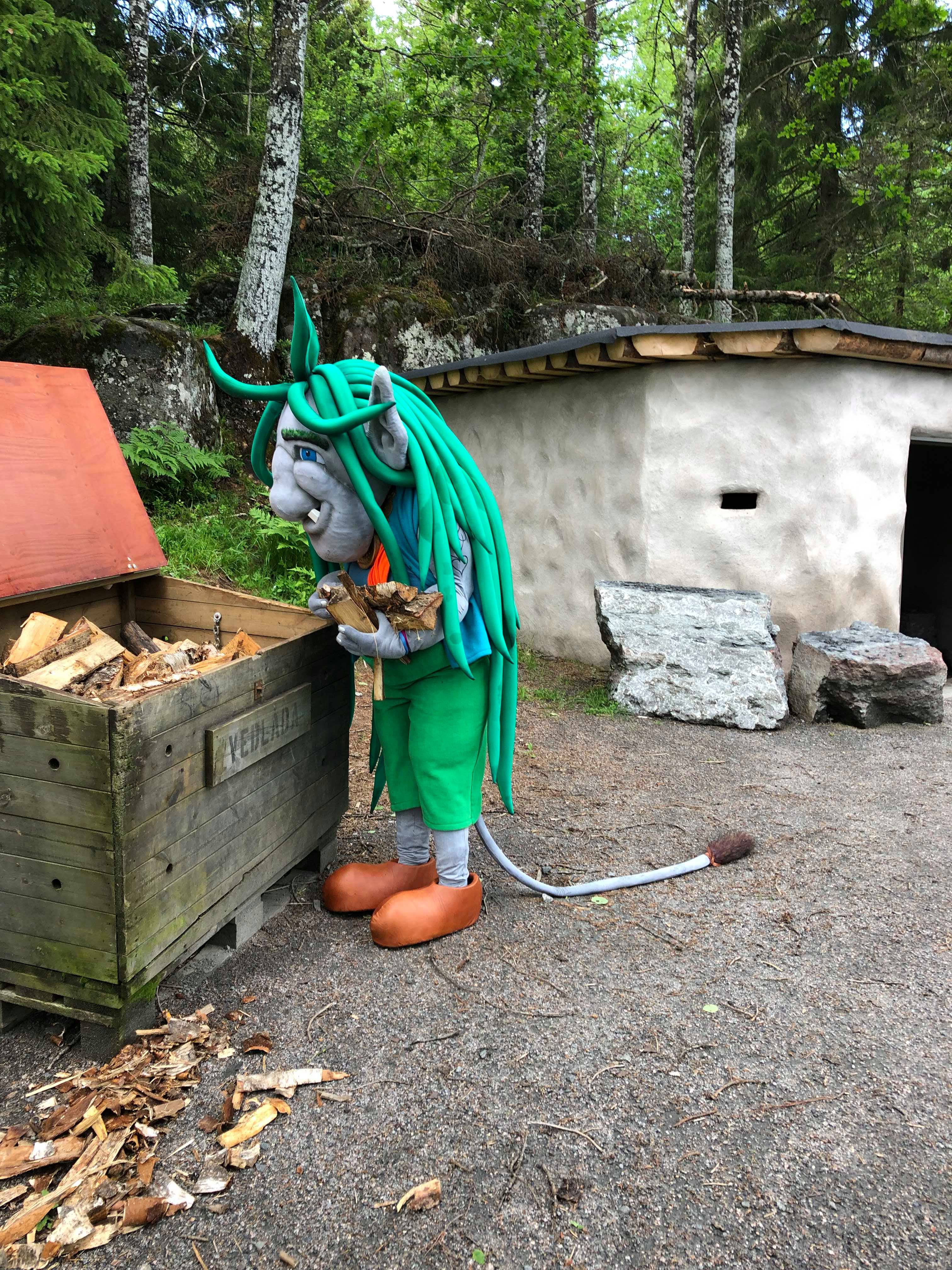 Ett stort troll med långt grönt hår står vid en låda och plockar ved. 