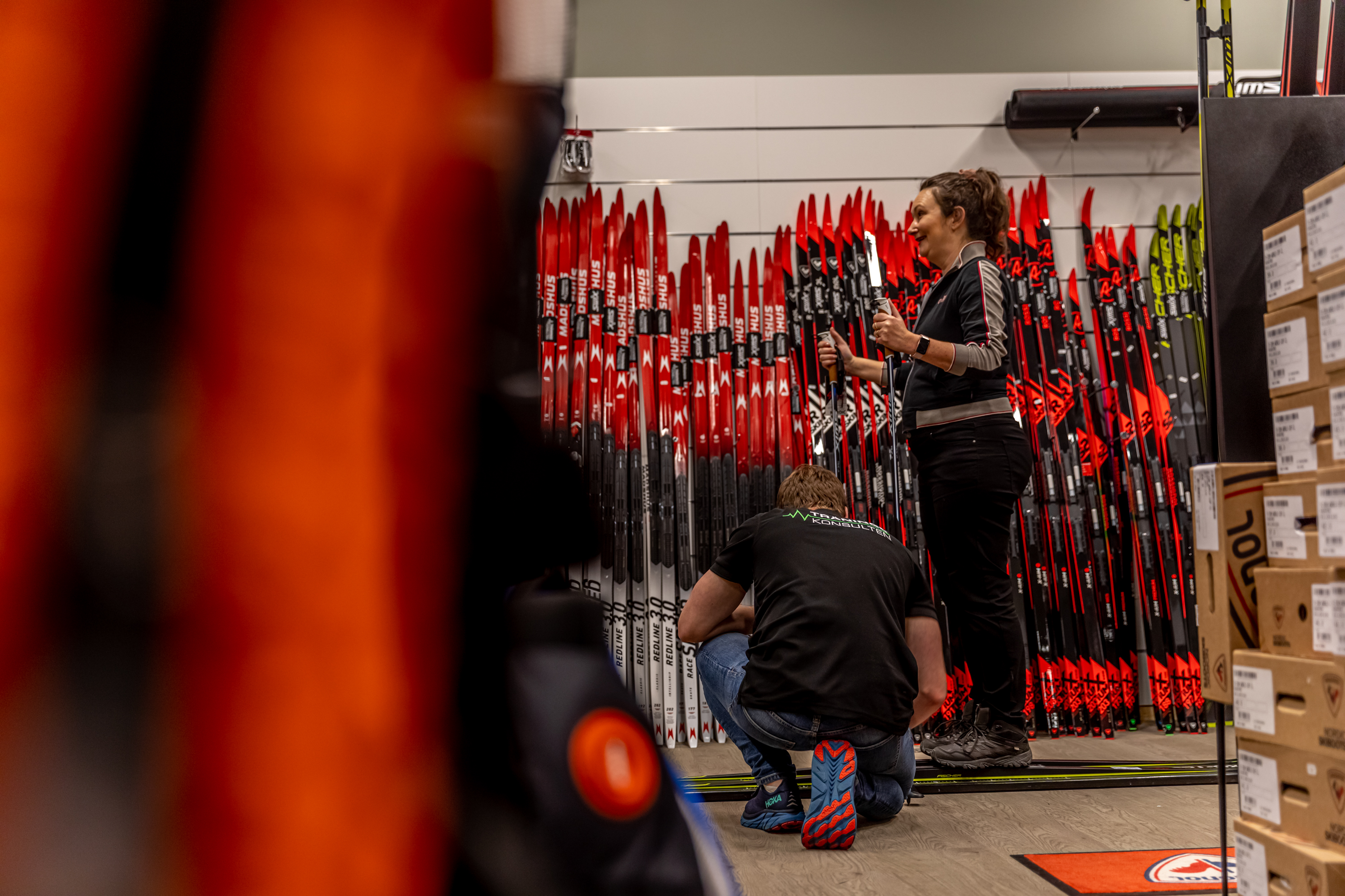 Kvinna står med längdskidor och stavar på sig i en sportbutik. En manlig expedit sitter på knä  och justerar hennes skidor.  