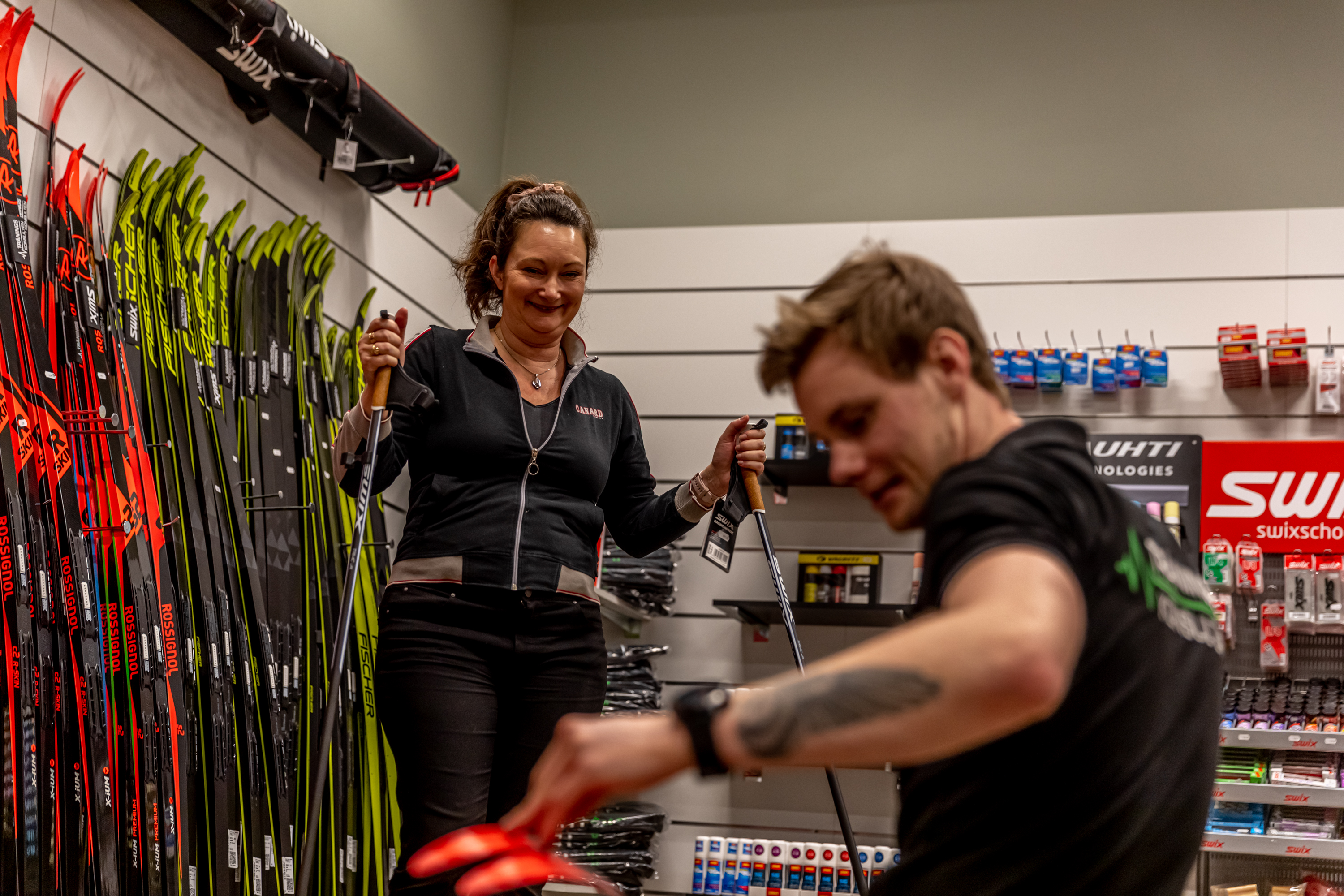 Kvinna står med stavar i en sportbutik. En manlig expedit sitter och plockar fram något till henne. I bakgrunden är en hel vägg med längdskidor. 