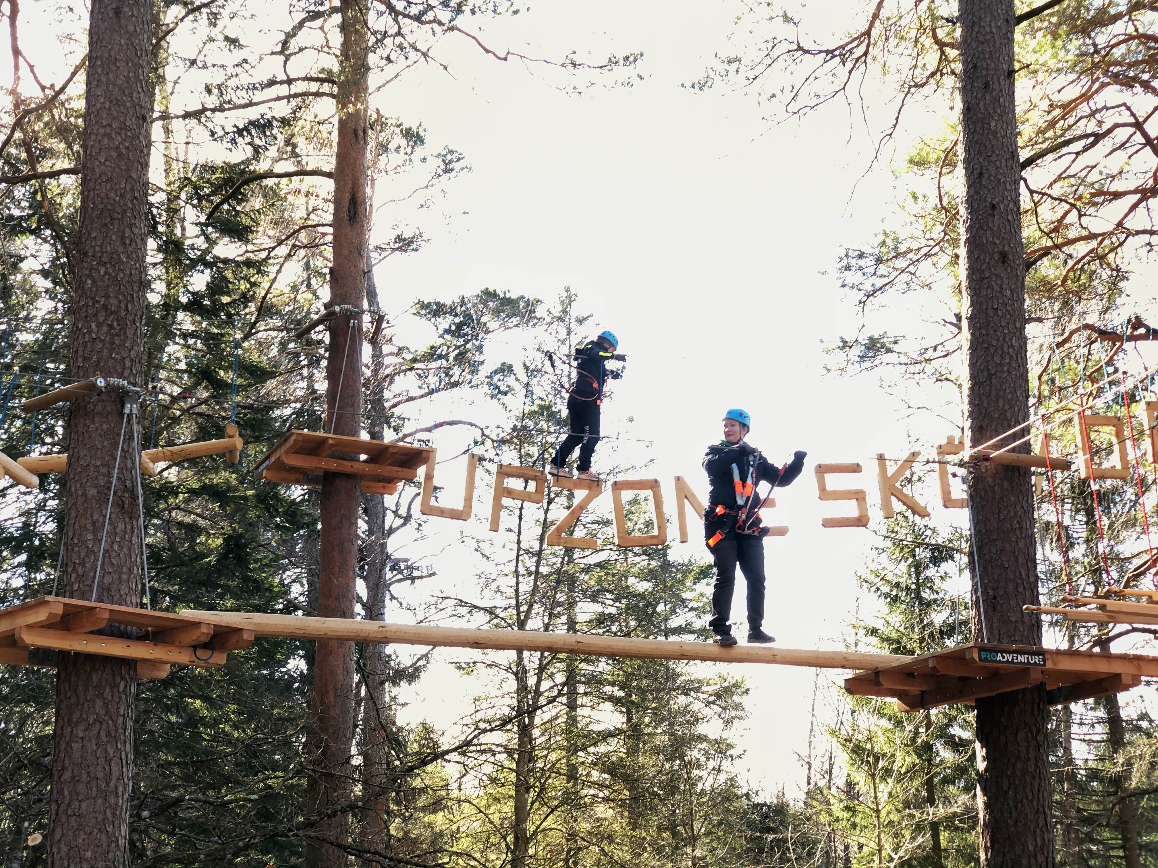Två personer balanserar på träspång respektive bokstäver som bildar ordet Upzone Skövde flera meter upp bland träden. 