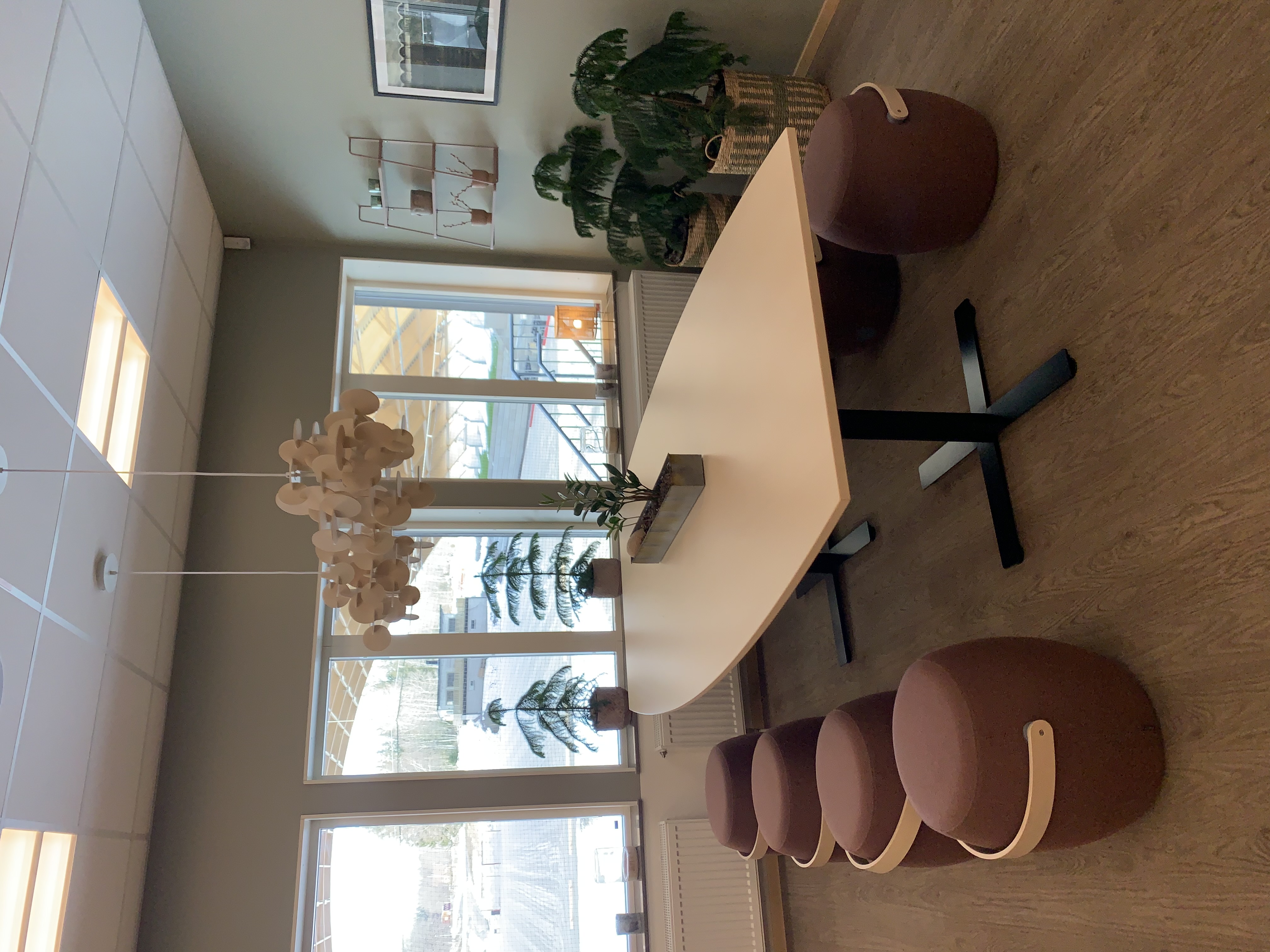 Konferensrum - ett stort ljus bord rosa textilpuffar på var sida om bordet. Två lampor gjord av cirklar i träfaner hänger ovanför bordet och växter i bakgrunden, i fönstret. 