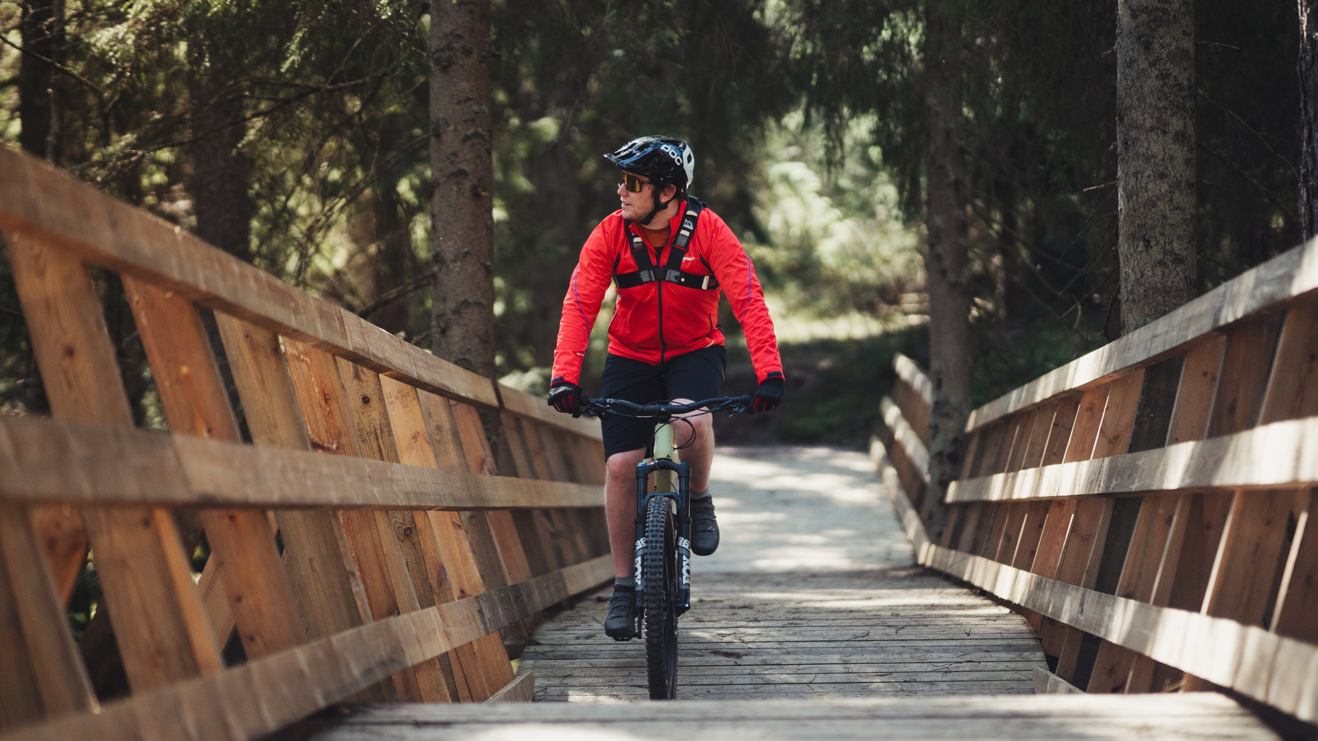 A man in a red jacket and black bike helmet is riding his mtb along a long ramp in the forest.