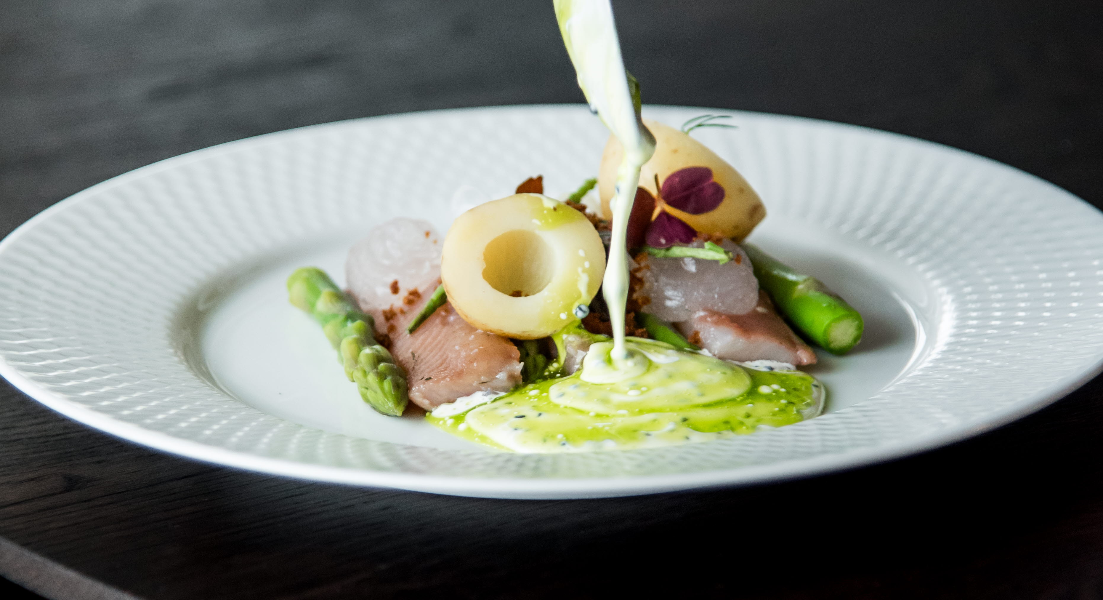 En närbild på en välkomponerad maträtt som ser väldigt lyxig ut. En person använder en sked för att lägga sås på tallriken. 