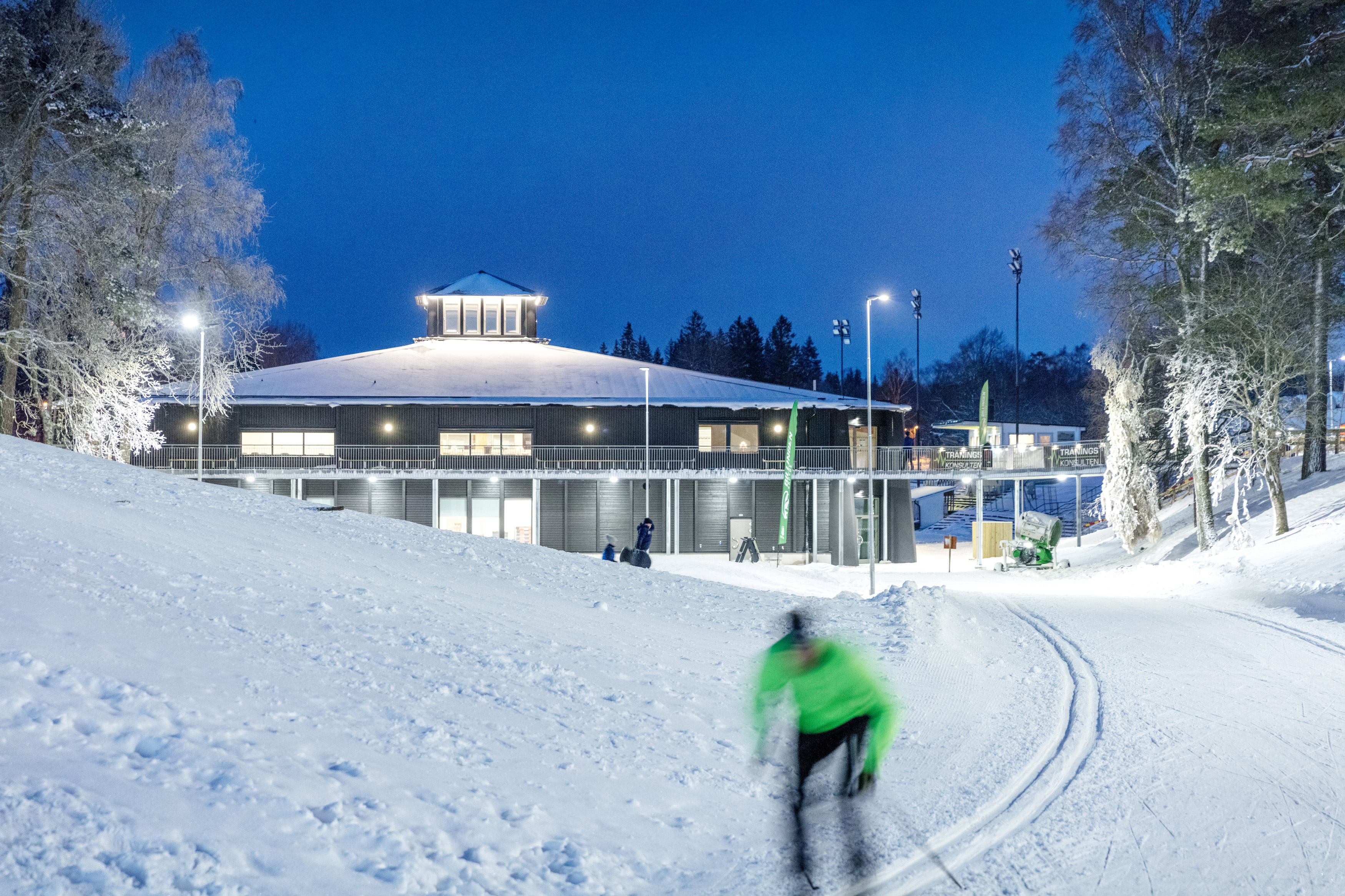 Skidåkare svischar förbi i spåren framför Billingecentret en vinterkväll på Billingen Skövde. 