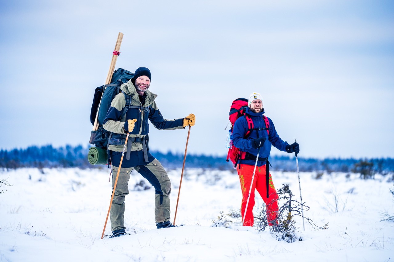 Två män står mitt i ett vinterlandskap i full vandrarutrustning med snöskor på fötterna. 