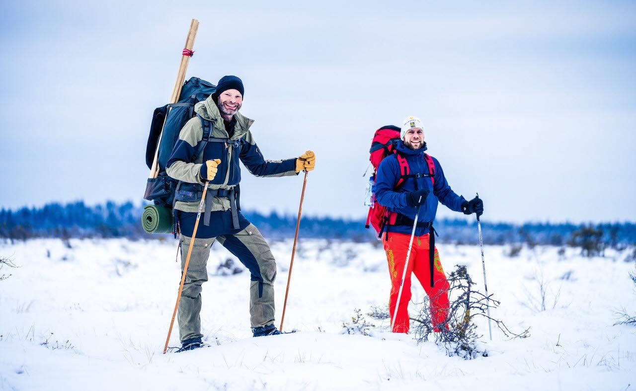 Två män står mitt i ett vinterlandskap i full vandrarutrustning med snöskor på fötterna. 