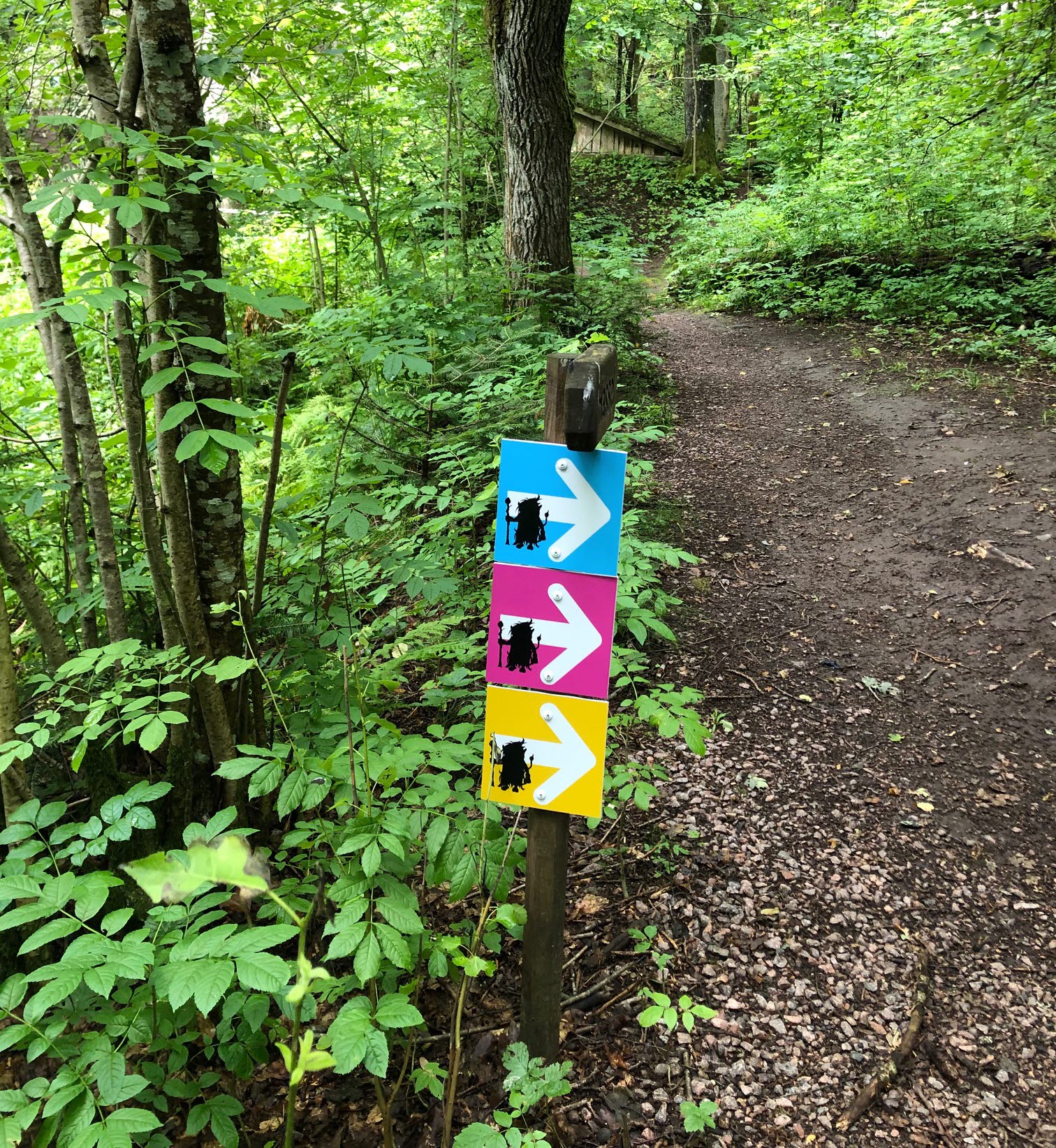 Skylt omgiven av grönska. Skylten föreställer tre pilar i blått, rosa och gult som ska visa vandrare vilken väg de ska gå. 