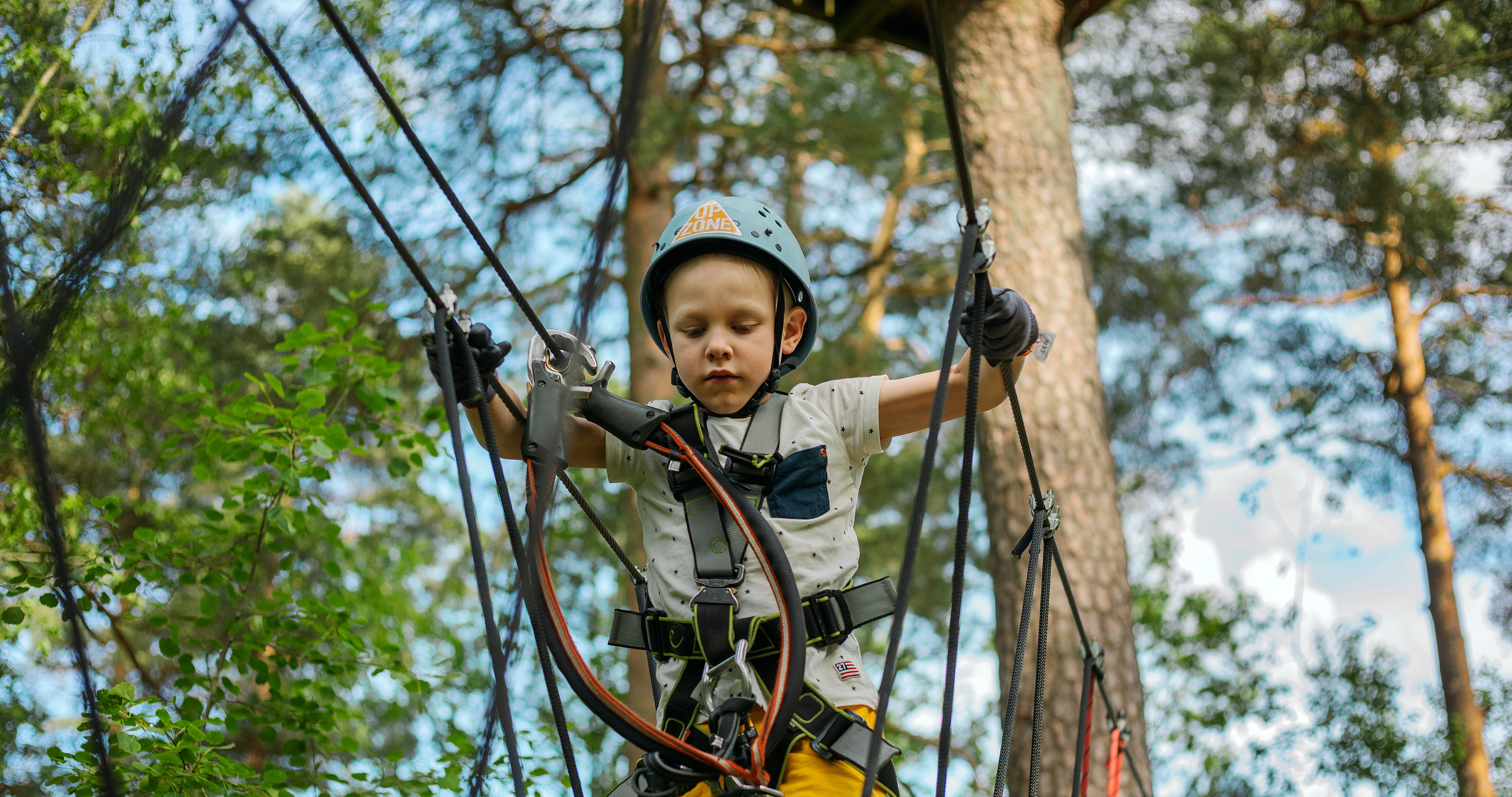 Ett barn balanserar på en lina högt uppe bland träden med sele på sig.