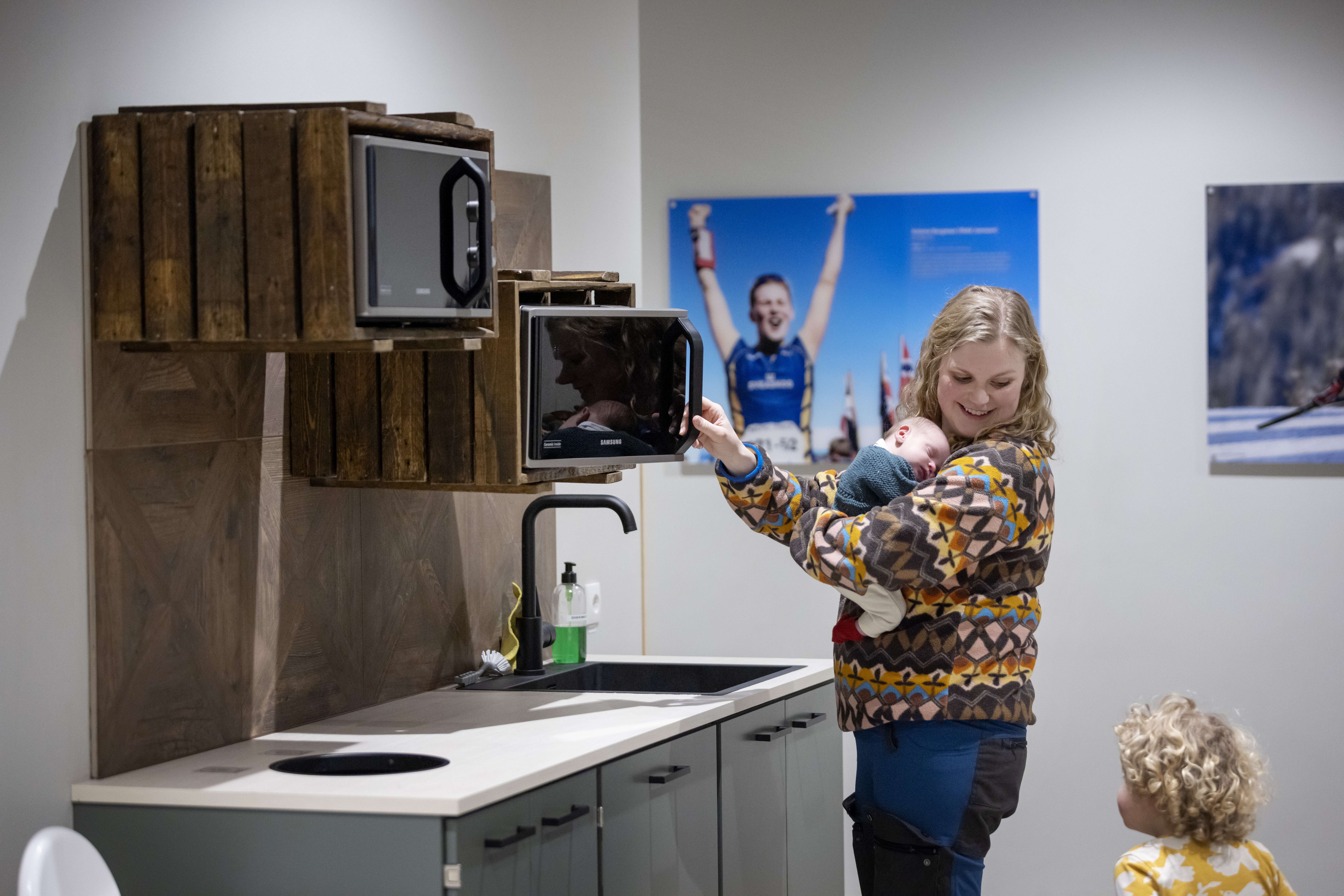 A mother is standing in a small kitchenette and has opened the microwave. On her arm she has a small baby. She looks over her shoulder and talks to her other child.