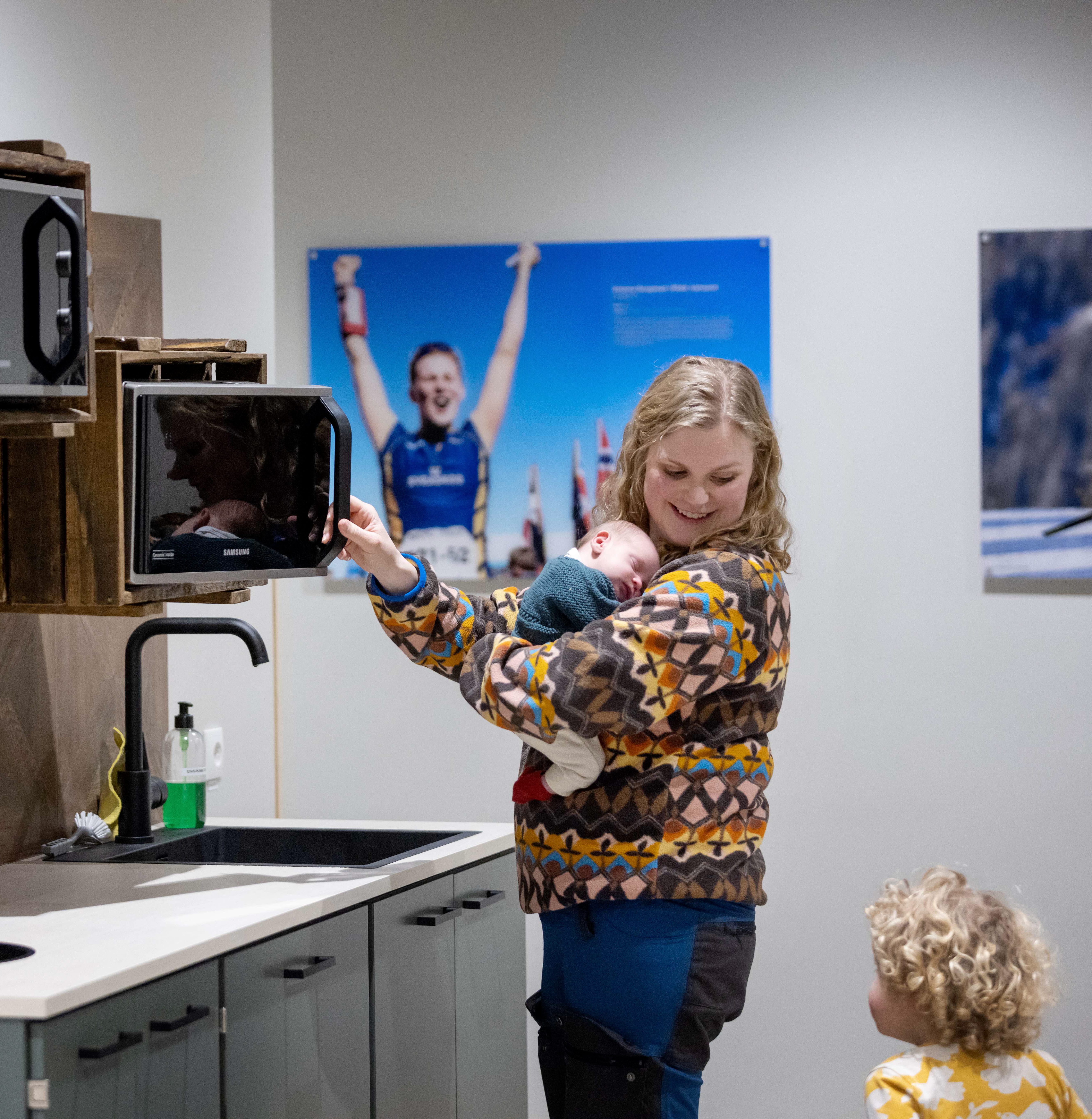 En mamma står i en litet pentry och har öppnat mikrovågsugnen. På armen har hon en liten bebis. Hon tittar över axeln och pratar med sitt andra barn.  