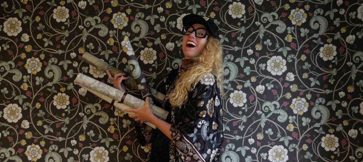 Kvinna som håller i tapetrullar framför en vägg i samma mönster. 