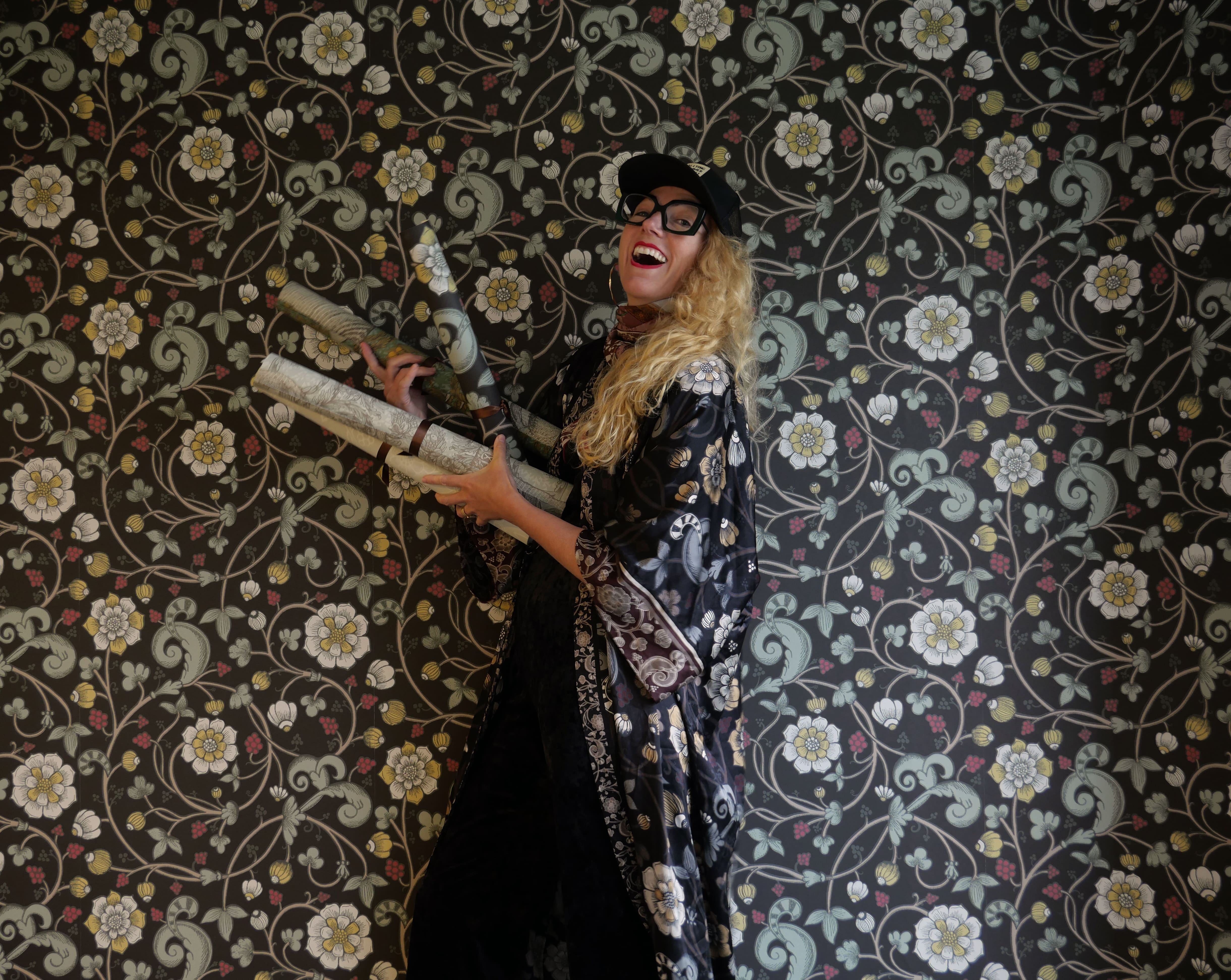 Kvinna med 2 tapetrullar i handen framför en vägg med blommiga tapeter.