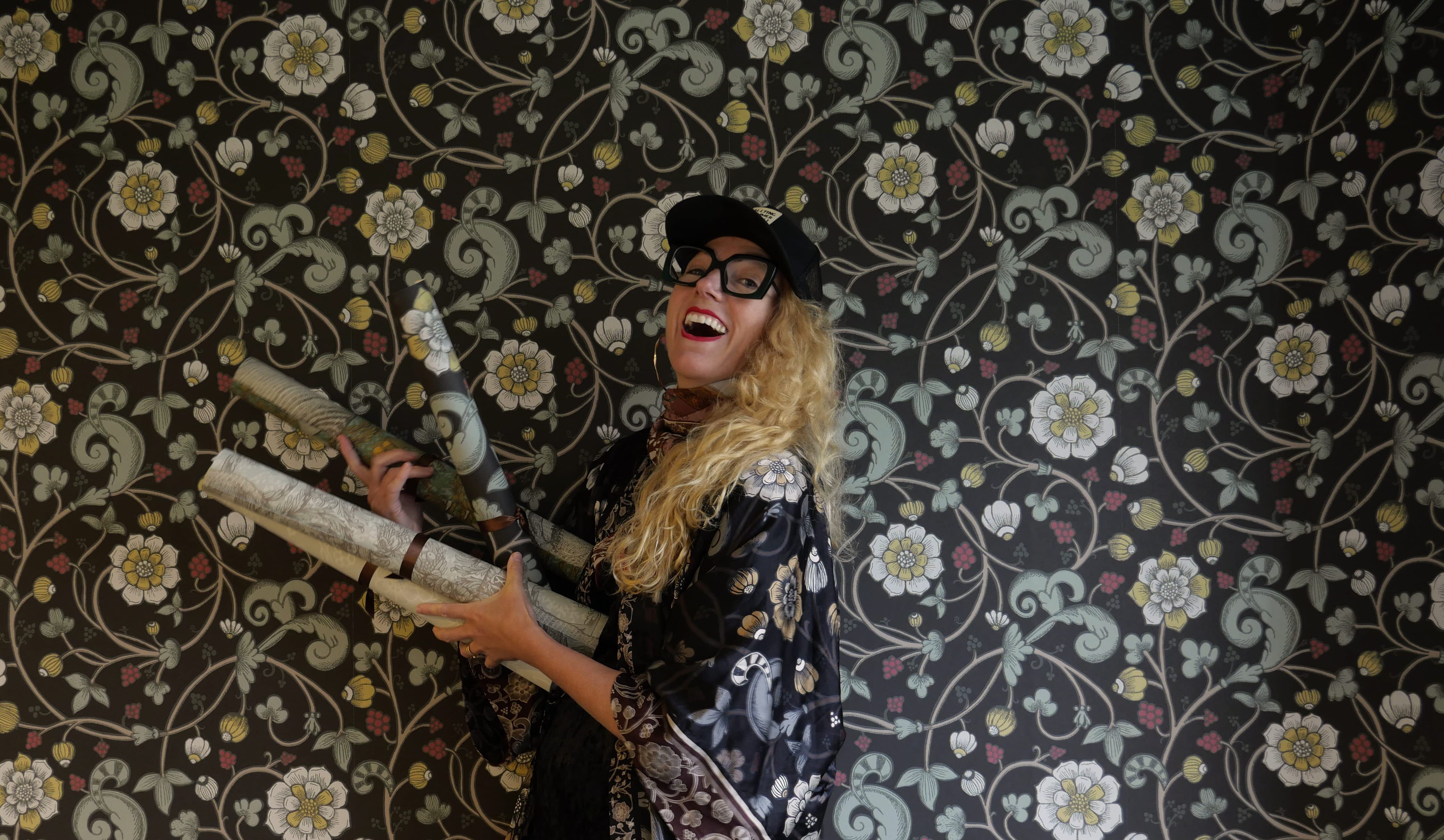 Kvinna med 2 tapetrullar i handen framför en vägg med blommiga tapeter.