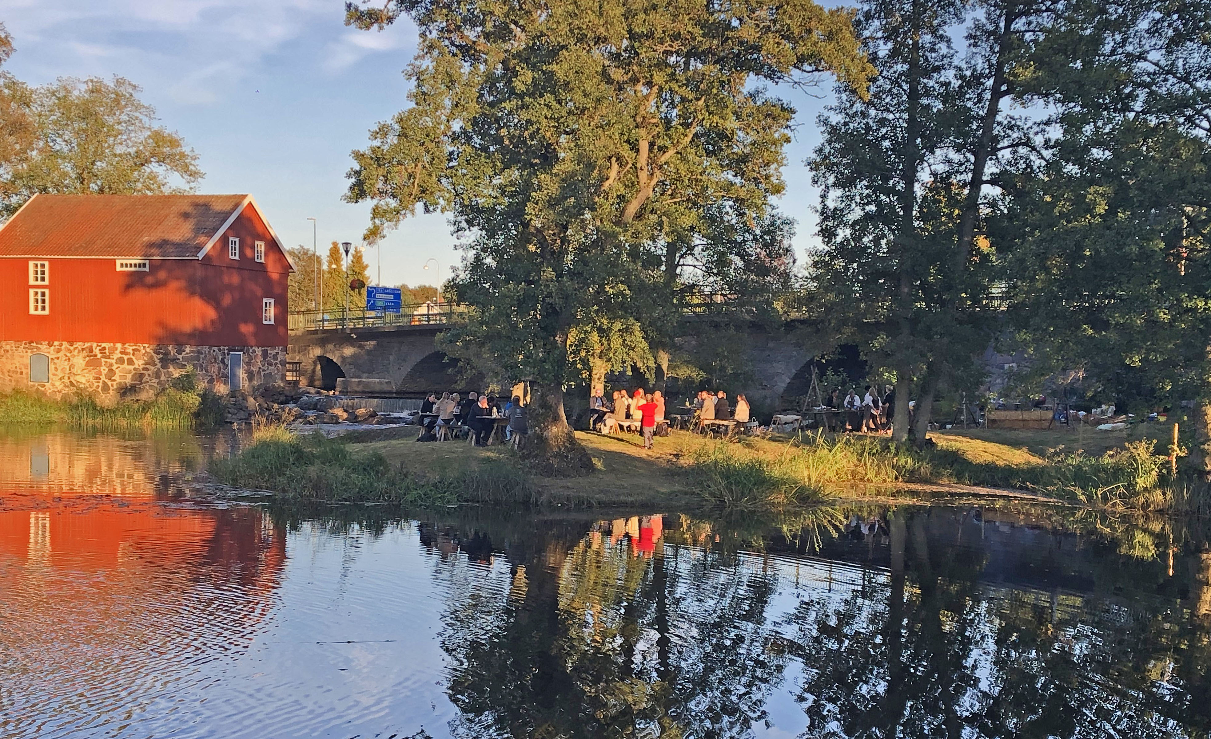 Ett rött hus till vänster i bilden med vatten framför och bredvid är det en sten bro och gröna träd. 