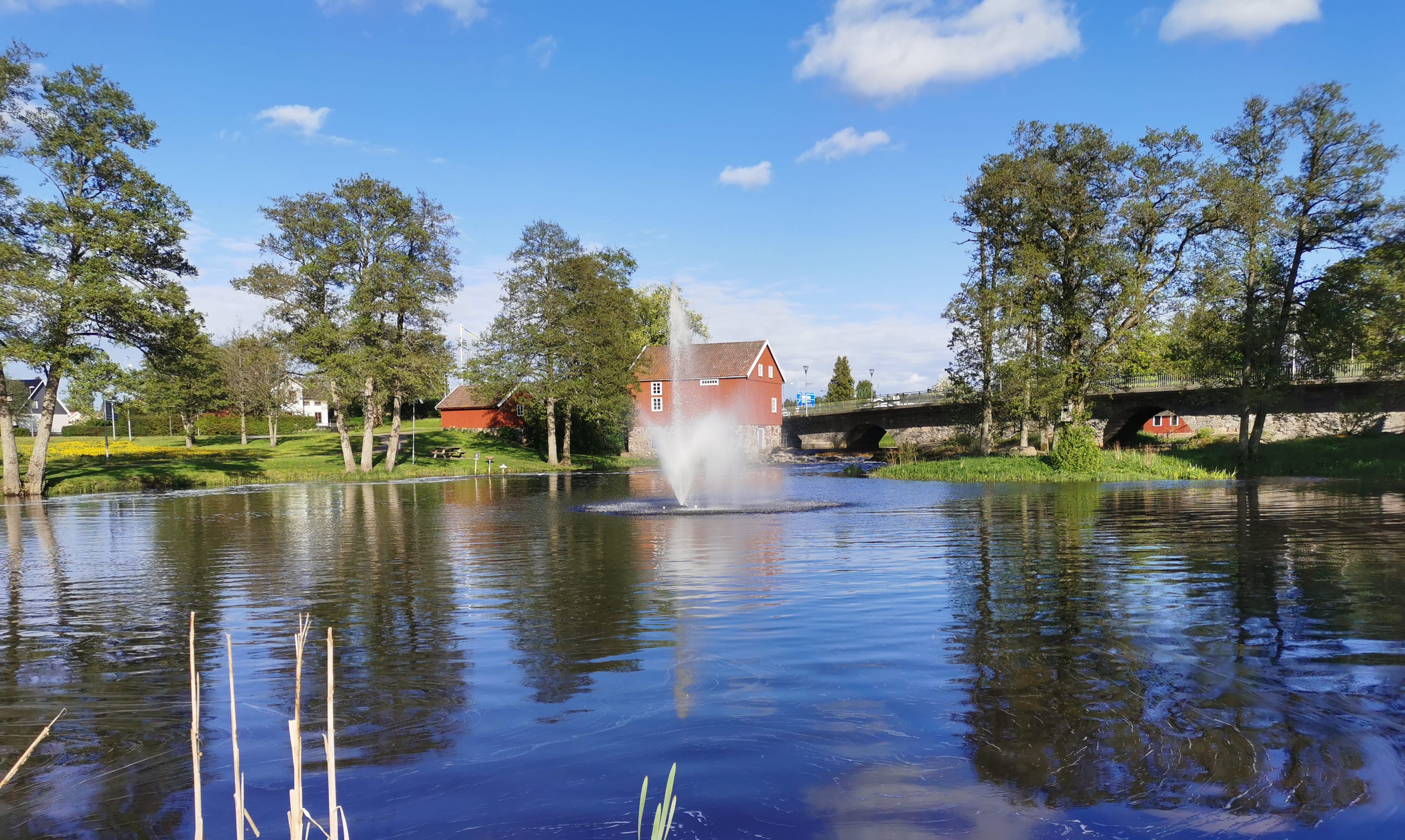 Vatten med en stor fontän i. Bakom sträcker sig en stor bro med en blå himmel ovanför. 
