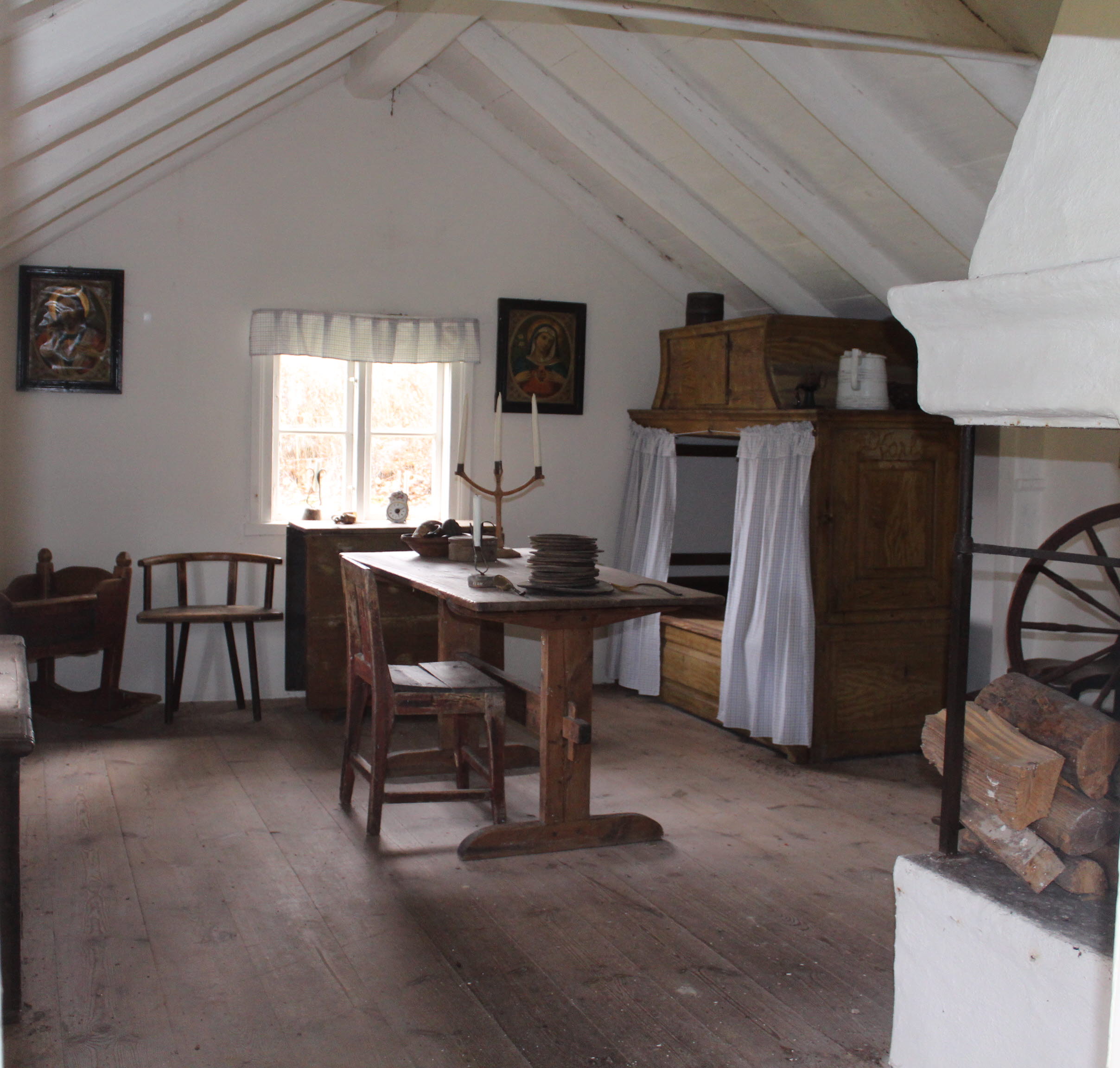 Inne i en hembygdsgård med ett gammalt träbord, trästolar och träsäng.