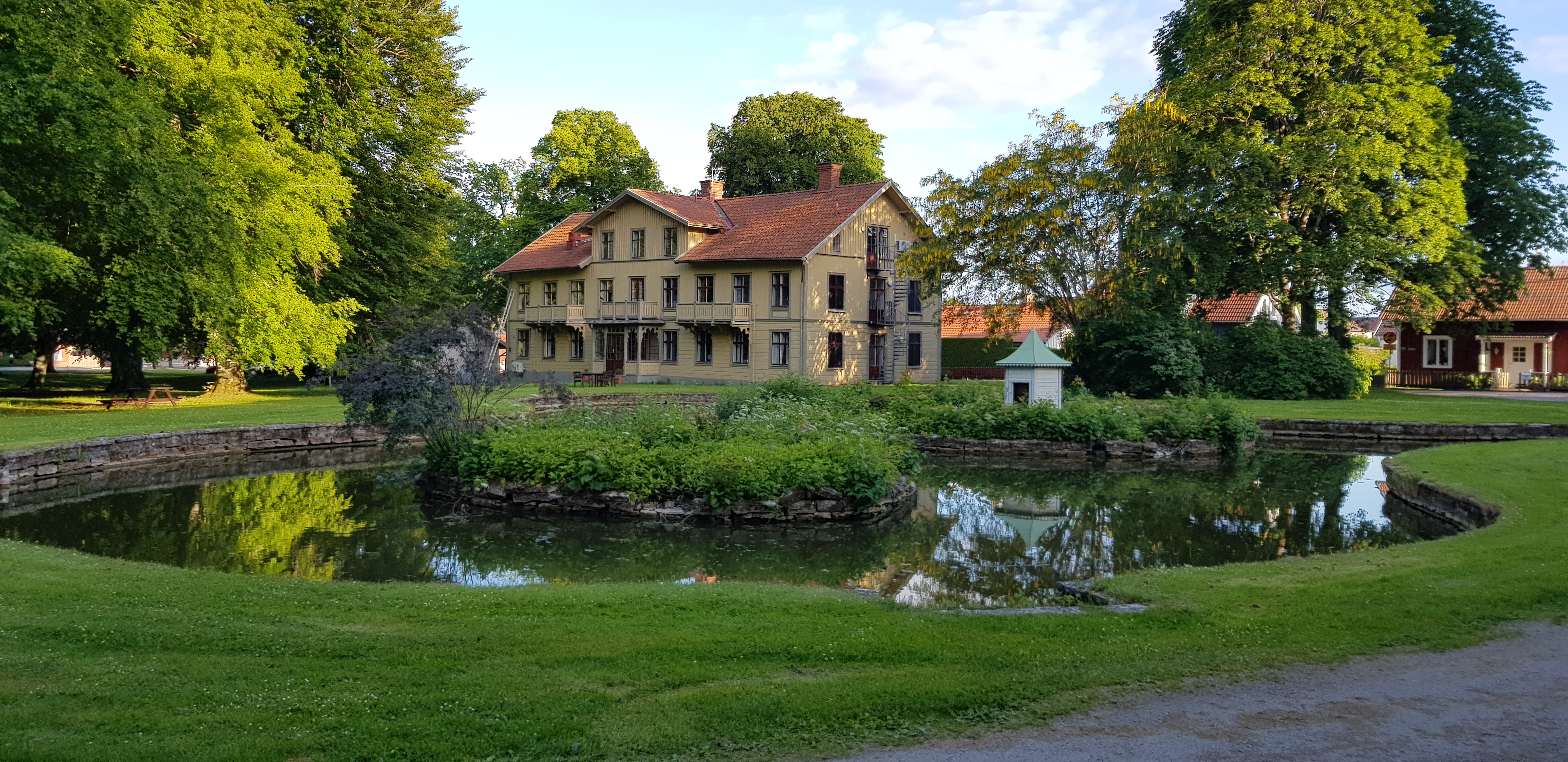 En damm och gräsmatta i förgrunden och ett gult stort hus i bakgrunden.