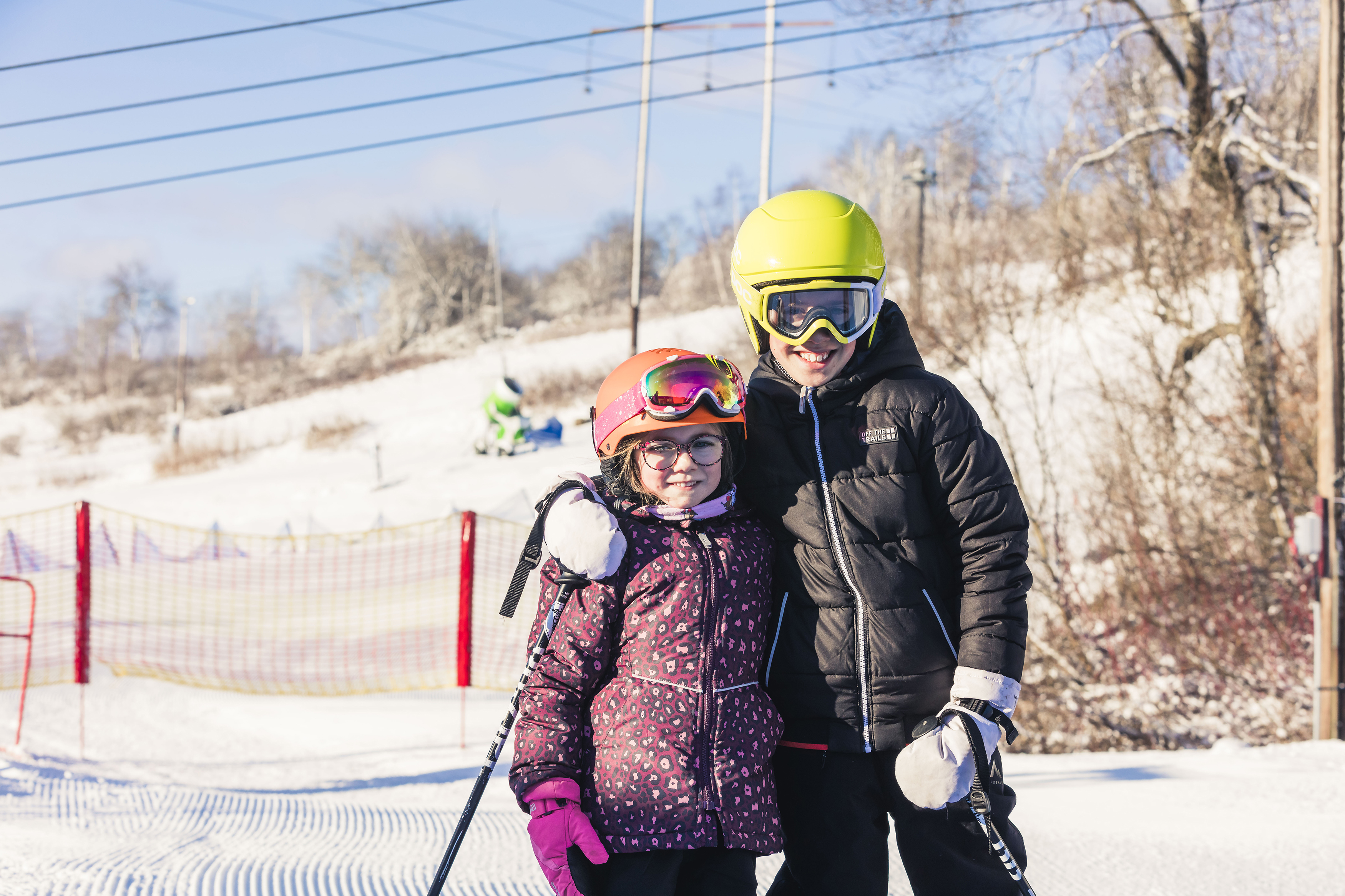 En pojke och en flicka står bredvid varandra med hjälmar och skidkläder på. Pojken har också skidglasögon på sig.