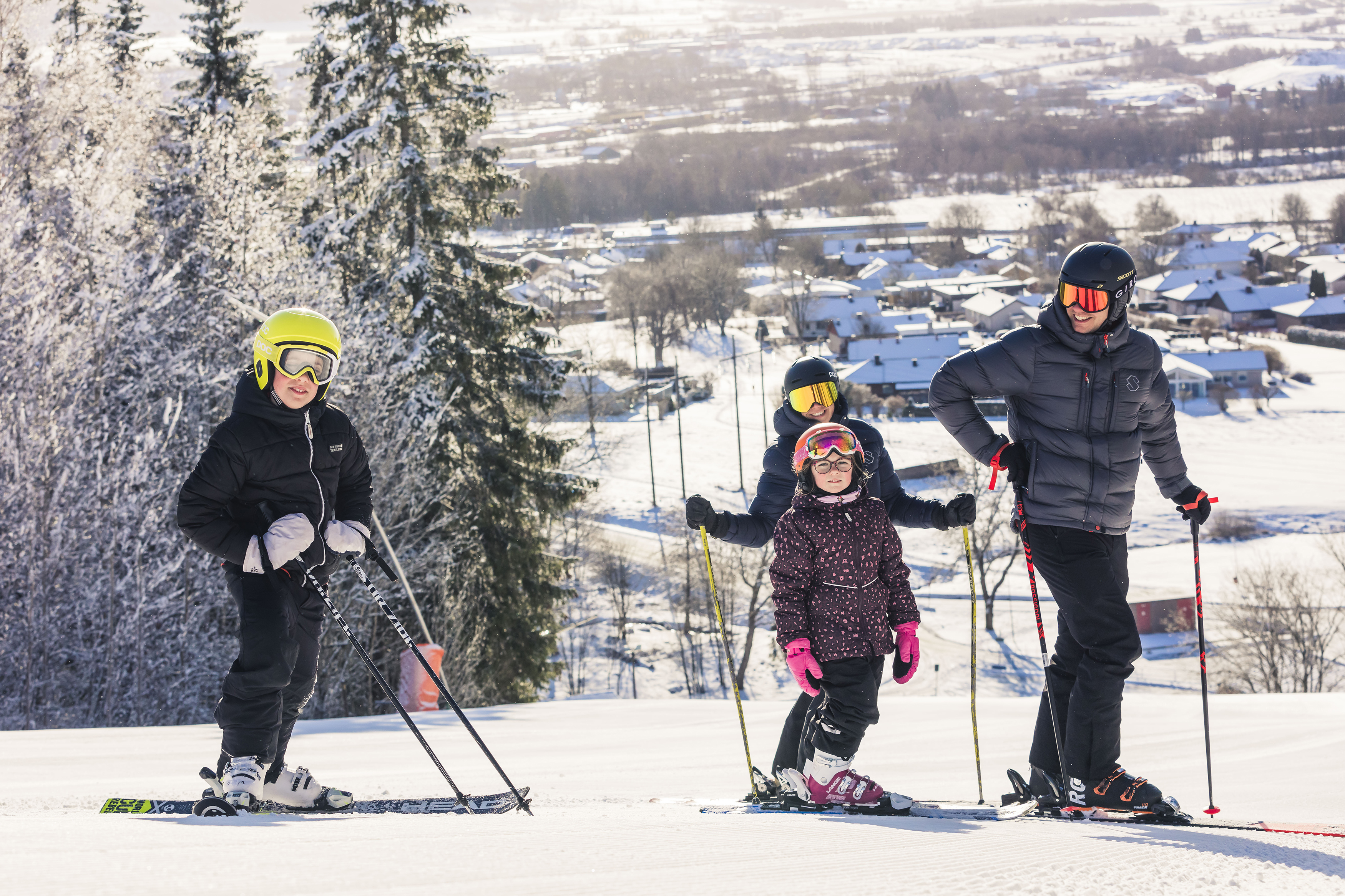 Två vuxna och två barn står i toppen på backen med sina skidor på. Solen skiner och man ser utsikten från berget bakom dem.