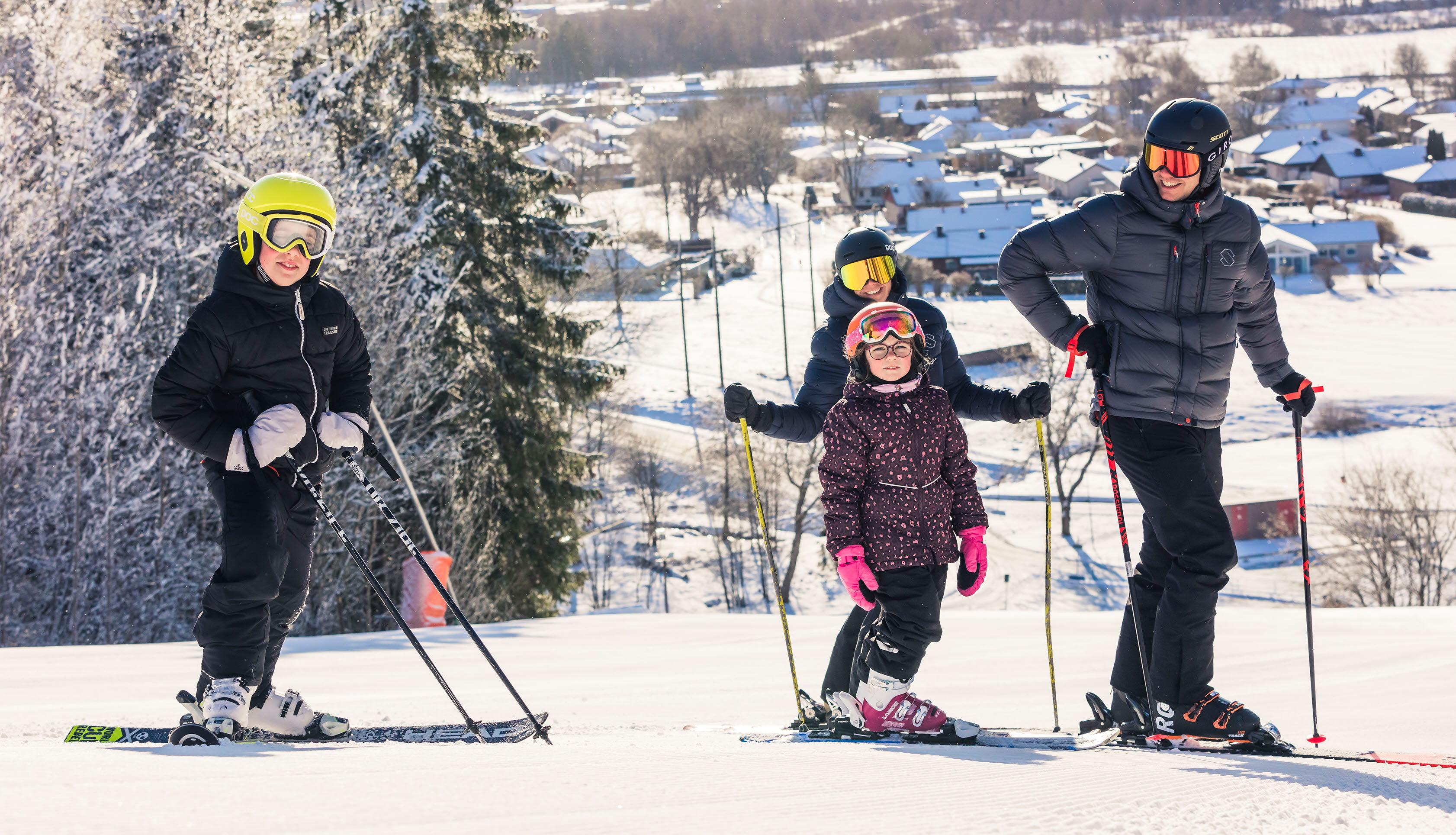 Två vuxna och två barn står i toppen på backen med sina skidor på. Solen skiner och man ser utsikten från berget bakom dem.