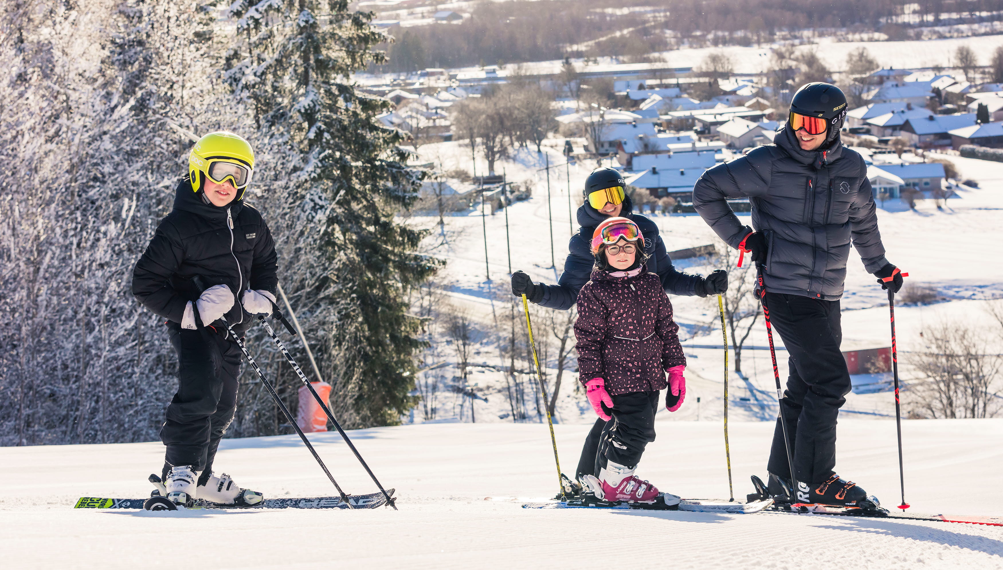 Two adults and two children stand at the top of the hill with their skis on. The sun is shining and you can see the view from the mountain behind them.