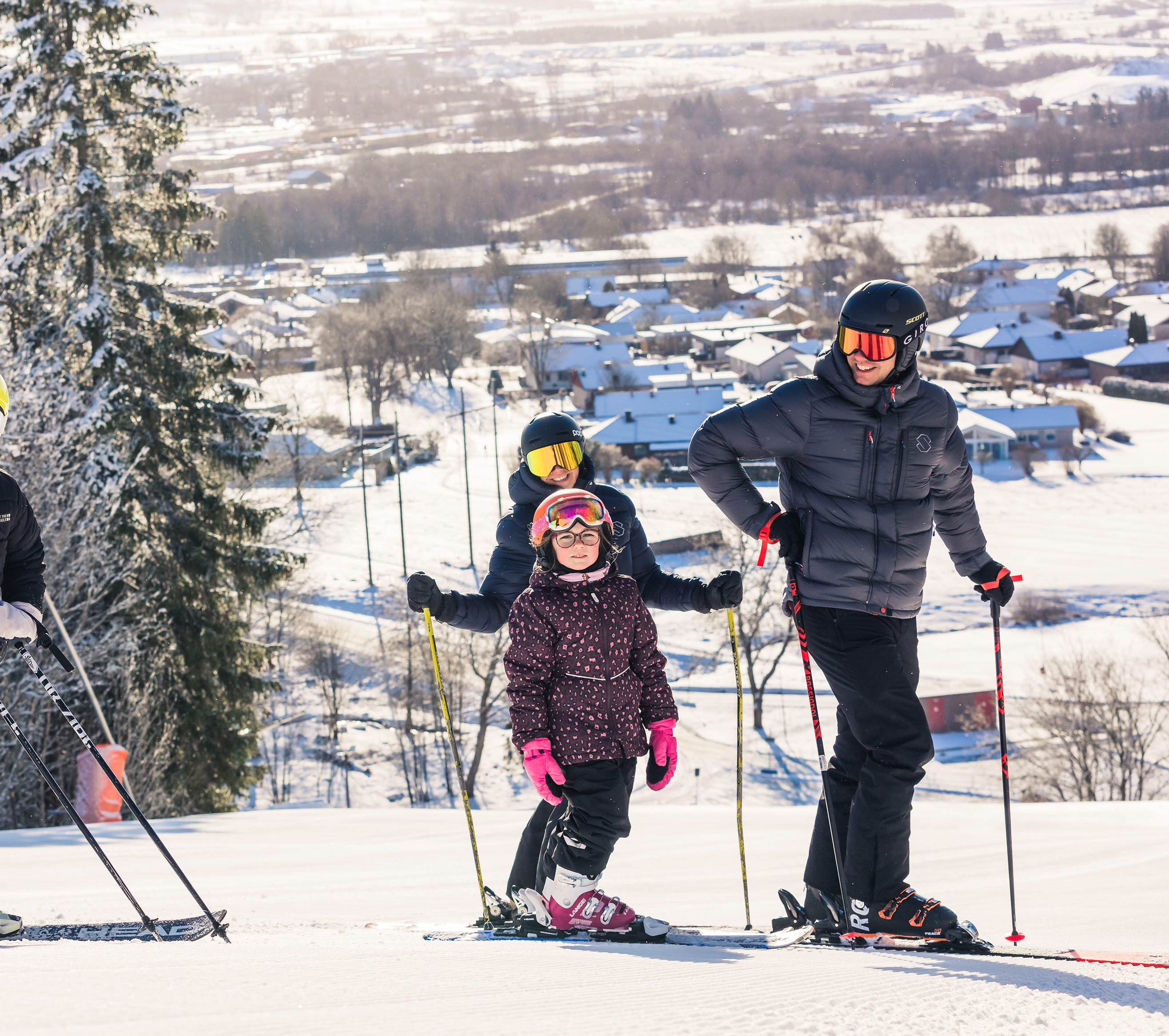 Två vuxna och två barn står i toppen på backen med sina skidor på. Solen skiner och man ser utsikten från berget bakom dem.