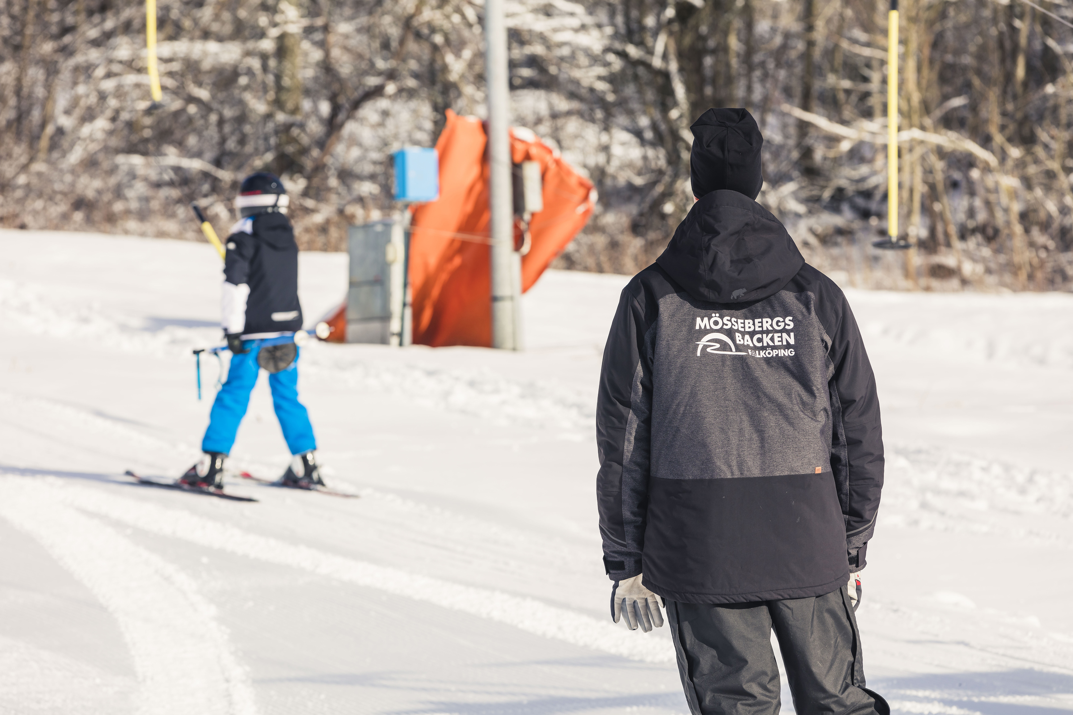 En liftvärd står med ryggen mot kameran med texten Mössebergsbacken på sin jacka. Ett barn är på väg upp i liften.