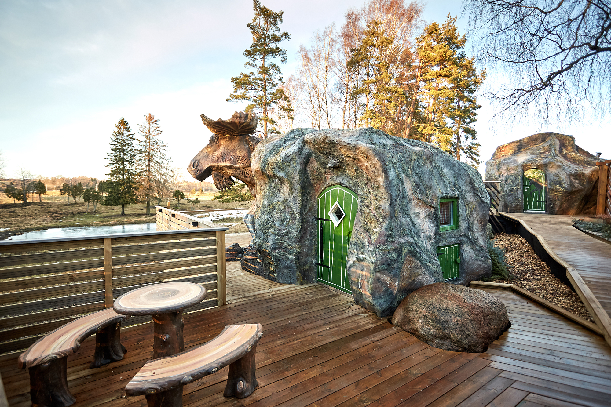 En stuga som ser ut som en älg. Gröna dörrar och bruna sittbänkar utanför.