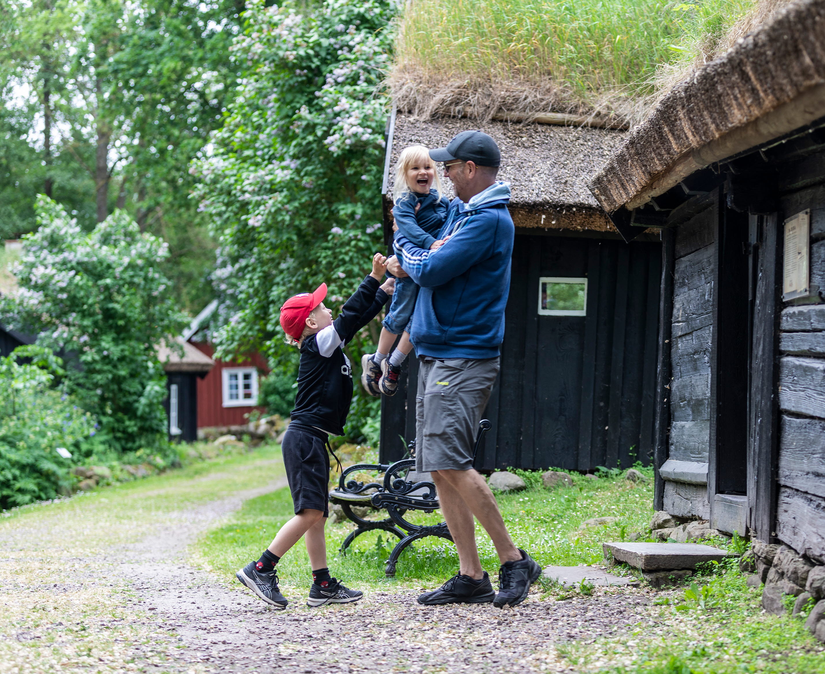Pappa med ett barn i famnen och annat barn står nedanför. Backstugorna i bakgrunden.